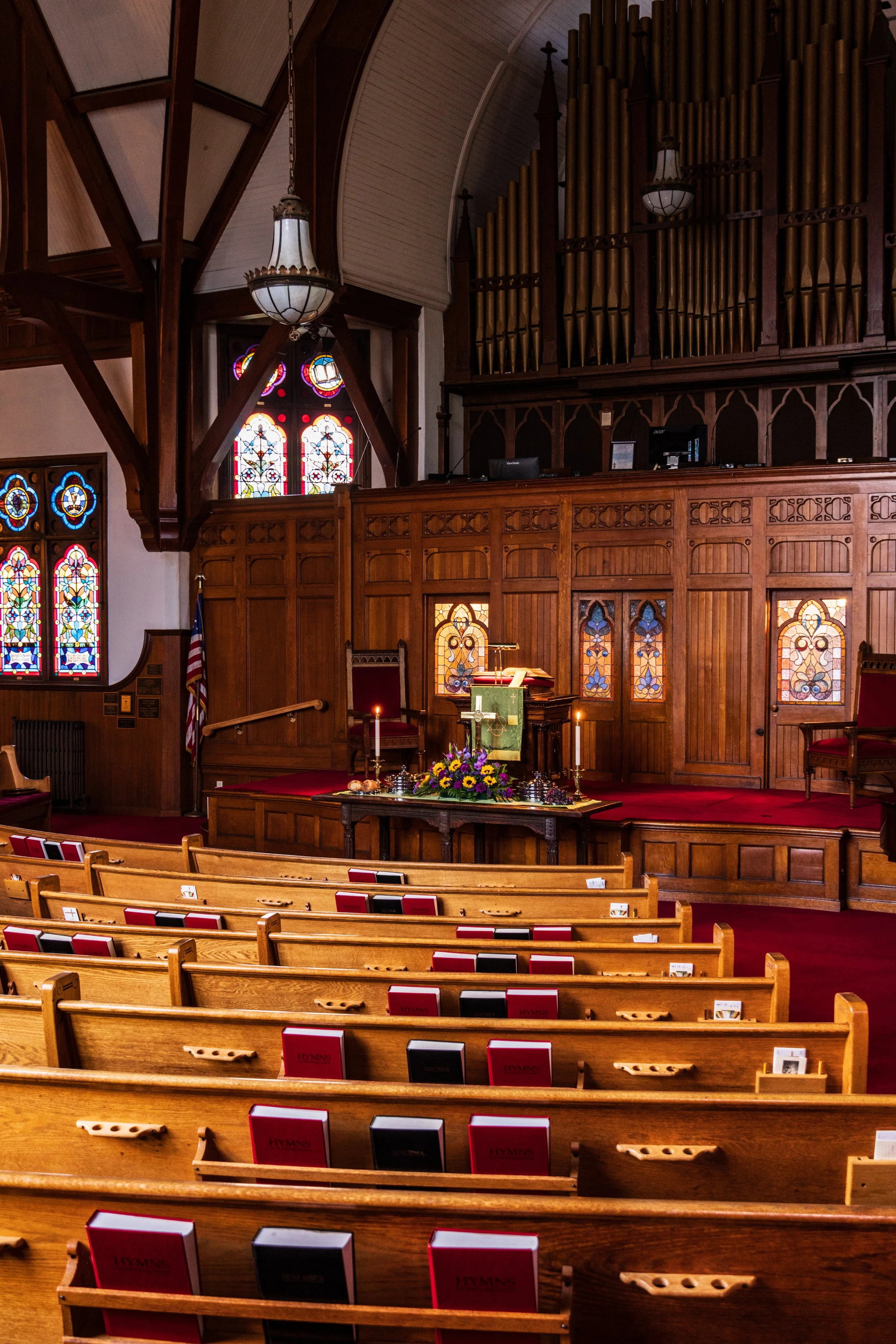 Interior view of a church with wooden pews, stained glass windows, an altar with a bouquet of flowers, candles, and hymn books, and a large pipe organ against the wall.