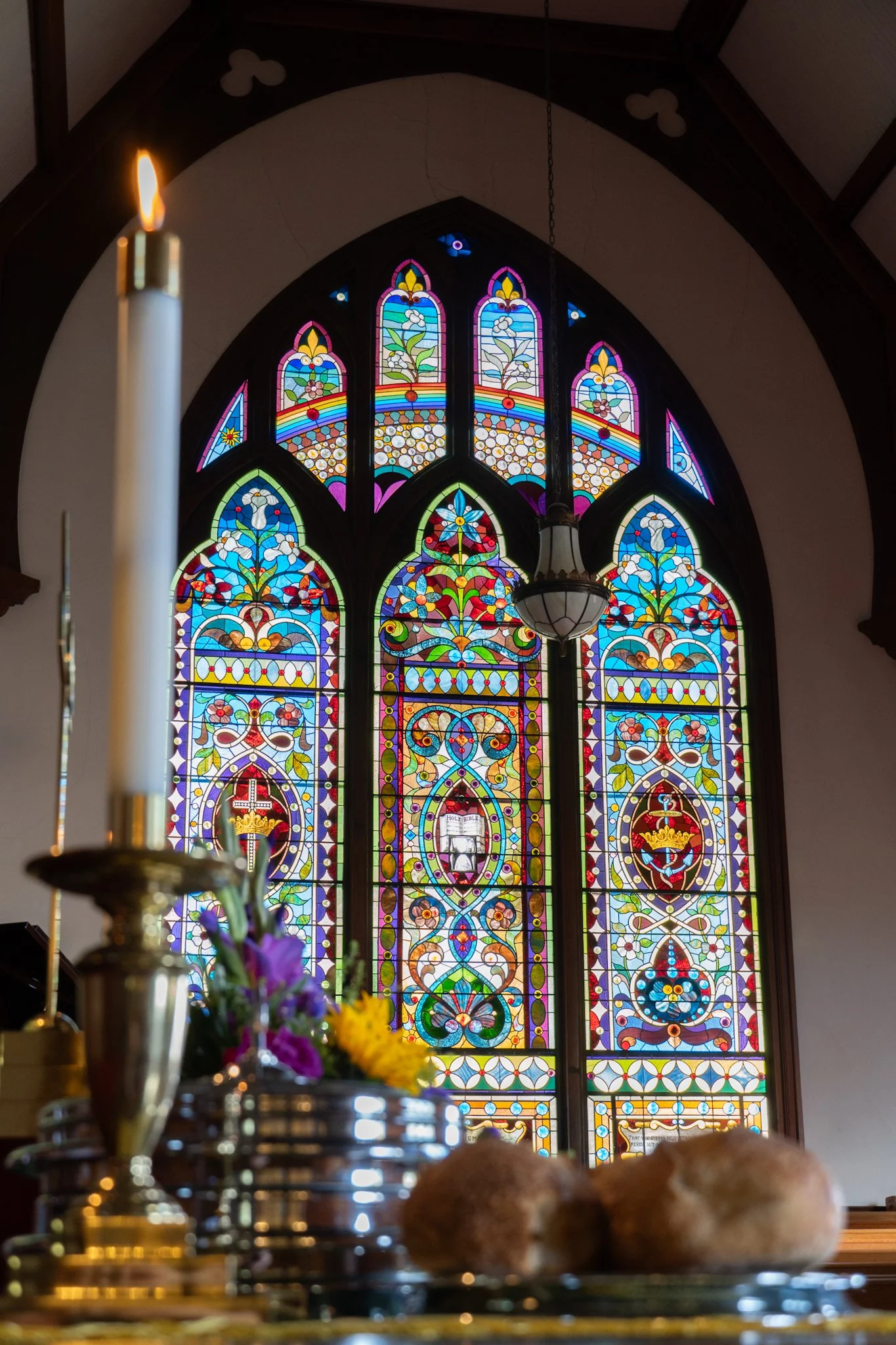 A church interior with colorful stained glass windows depicting floral and symbolic patterns. In the foreground, there are candles and flowers on a table.