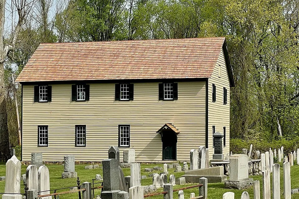 A large, two-story beige house with black shutters and a red-tiled roof, surrounded by a graveyard with various tombstones and monuments in the foreground, and green trees in the background.
