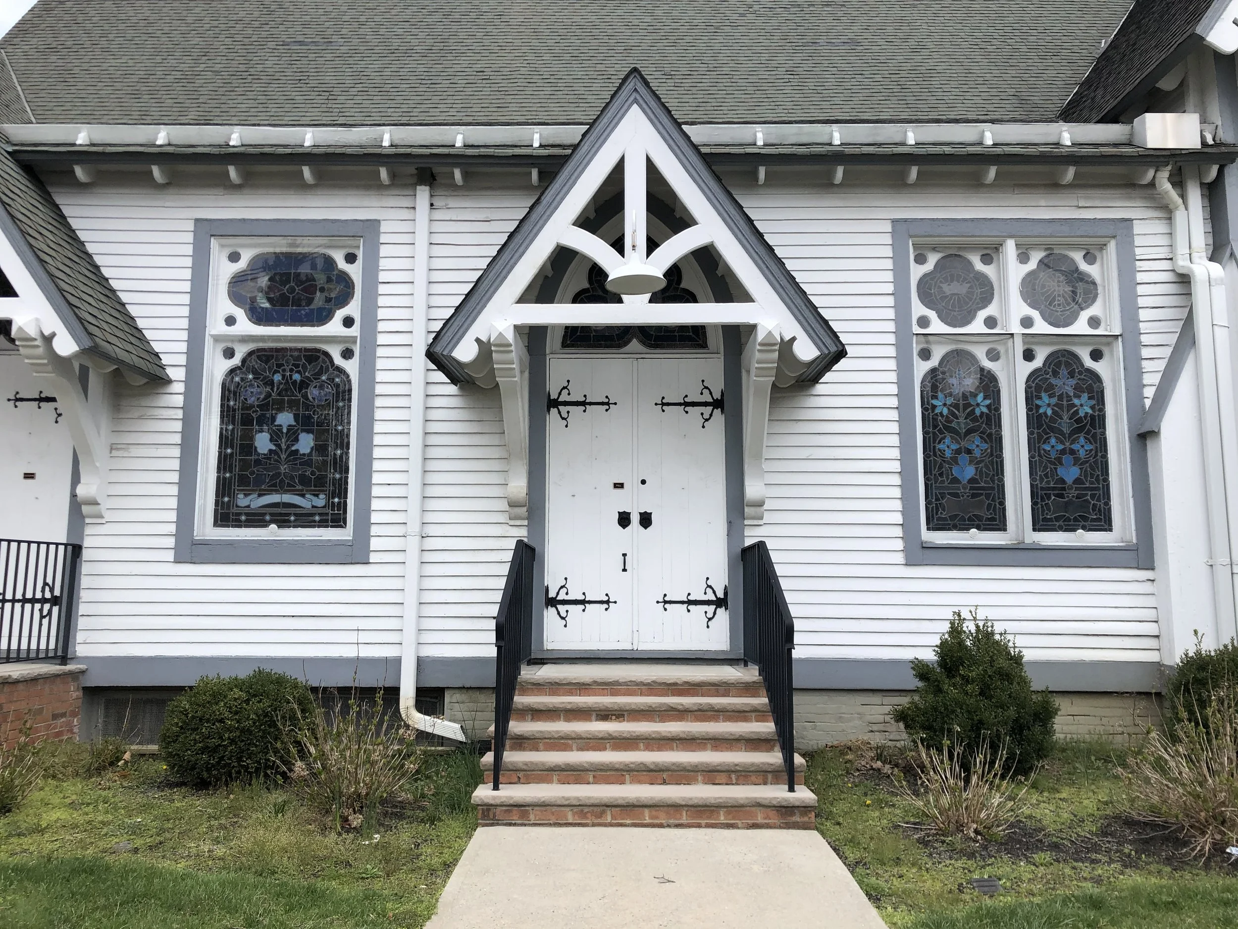 White wooden house with pointed gable roof and Gothic-style stained glass windows, black metal railing along brick steps, and garden bushes in the front.