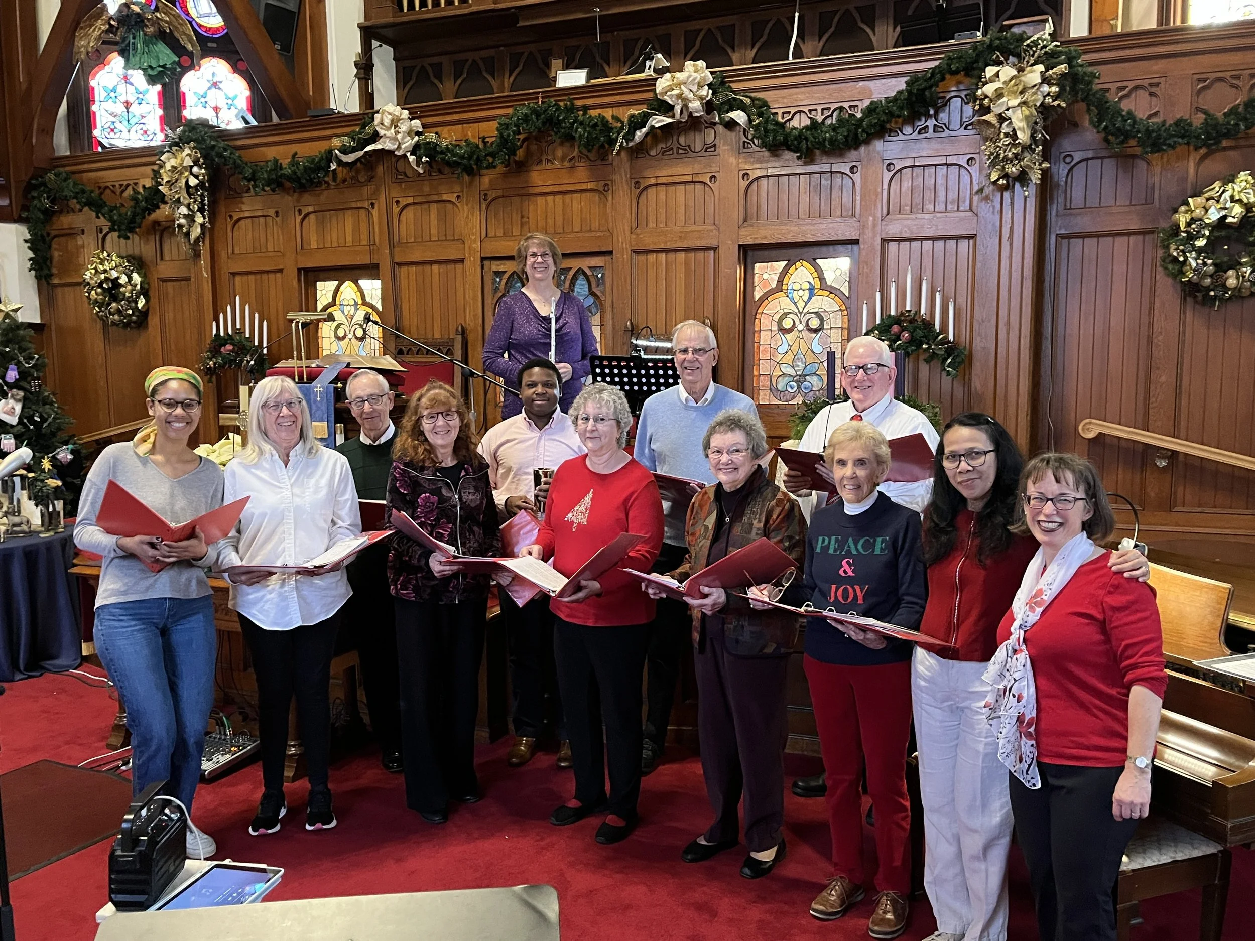 A group of people standing on a red carpet in a church decorated for Christmas, with stained glass windows and festive wreaths on the walls, some holding songbooks and singing.
