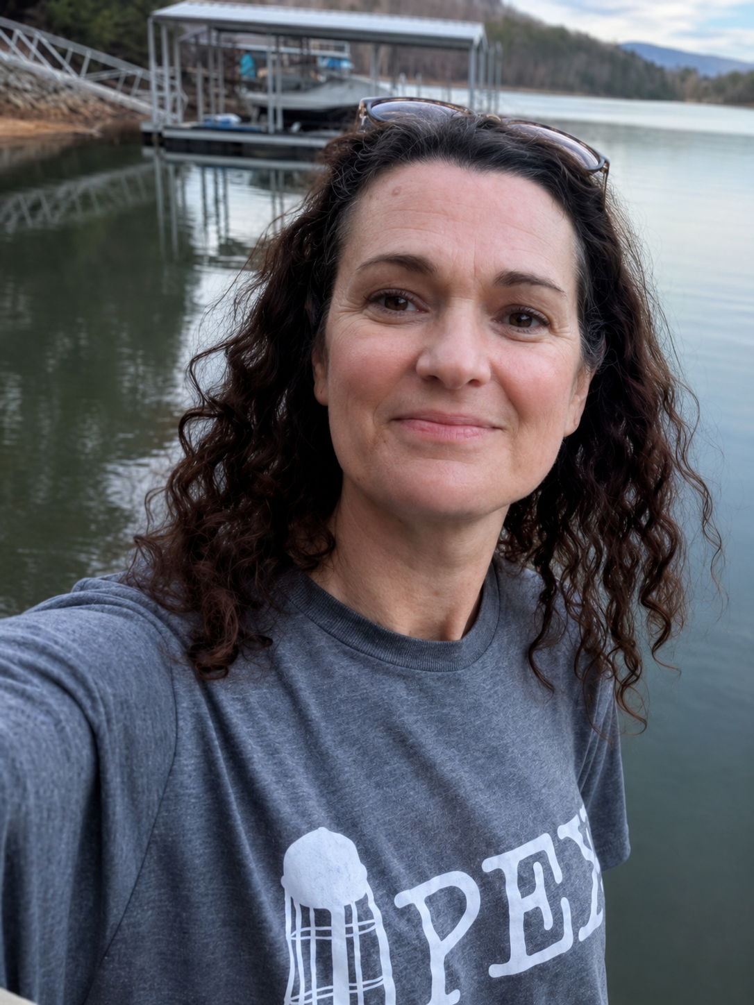 A woman with curly dark hair taking a selfie near a lake with a boat dock in the background.