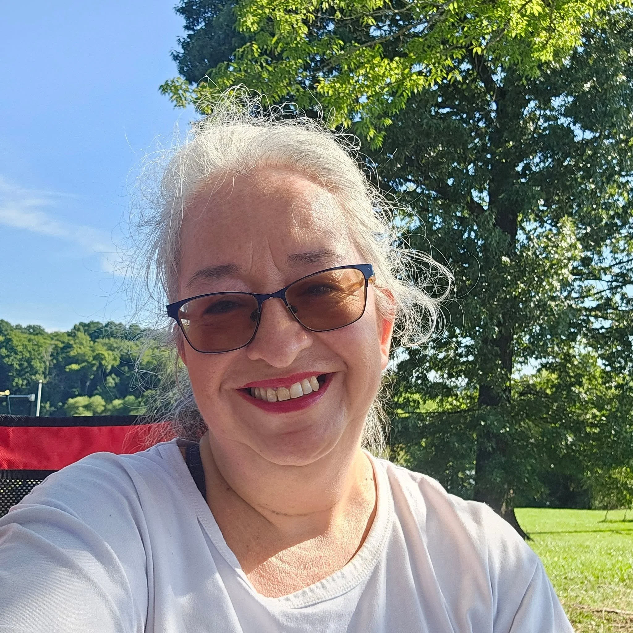 A woman with white hair and glasses smiling outdoors on a sunny day, with trees and a blue sky in the background.