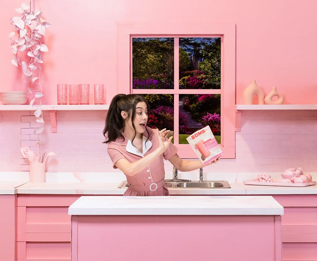 A woman in a pink kitchen holding a box of Bomb Bomba with an expression of surprise. The kitchen is decorated in shades of pink with shelves, vases, and utensils, and has a window showing a colorful garden outside.