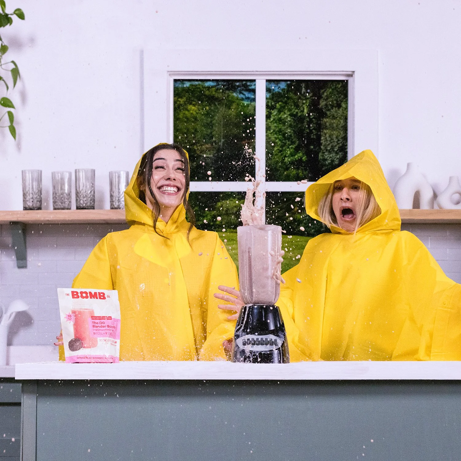Two women in yellow raincoats are standing in a kitchen, making a smoothie in a blender. The woman on the left is smiling broadly, while the woman on the right has a surprised expression. They are wearing hooded rain jackets, and splashes of smoothie are seen mid-air. There is a packet labeled "The BOMB" on the counter.