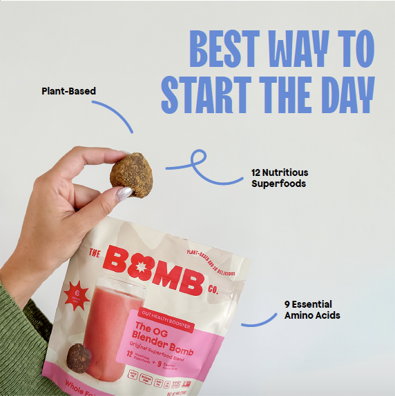 Hand holding a plant-based, nutrient-rich snack ball above a labeled package of The OG Blender Bomb, with large blue text that says 'Best Way to Start the Day' and notes indicating the snack contains 12 nutritious superfoods and 9 essential amino acids.