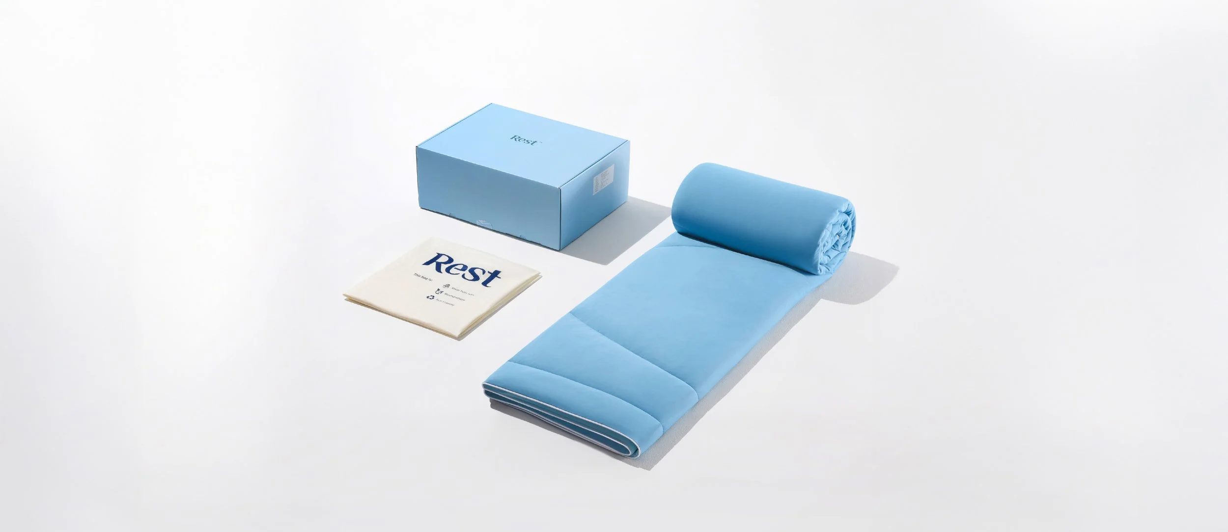 A blue portable massage bed with a rolled-up face cradle, a blue box labeled 'Reser,' and a cloth with 'Rest' printed on it, arranged on a white surface.