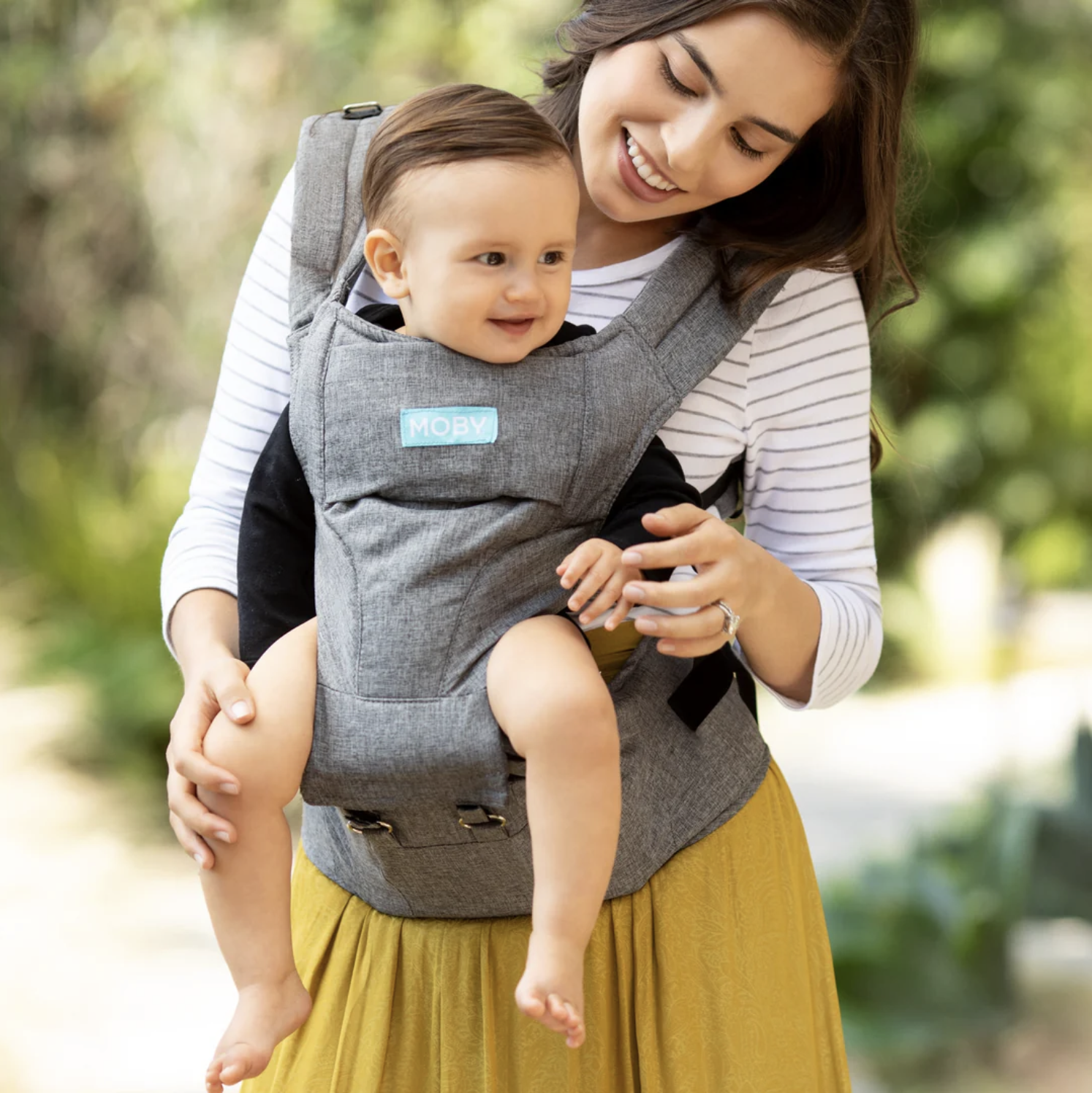A woman is smiling and carrying a young child in a gray Moby baby carrier outdoors. The child is looking to the side with a happy expression. The background is blurred greenery.