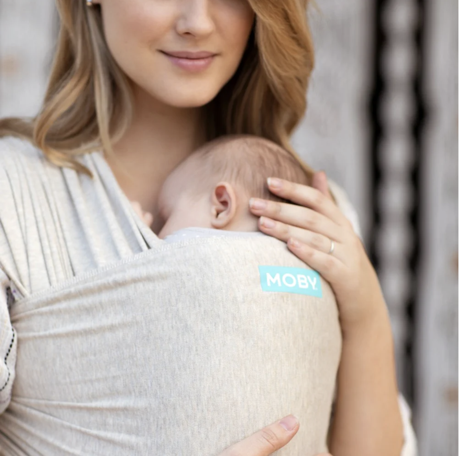 A woman with red hair holding a baby close to her chest in a beige carrier labeled 'MOBY', with the baby's head resting on her chest and her hand gently supporting the baby's head.