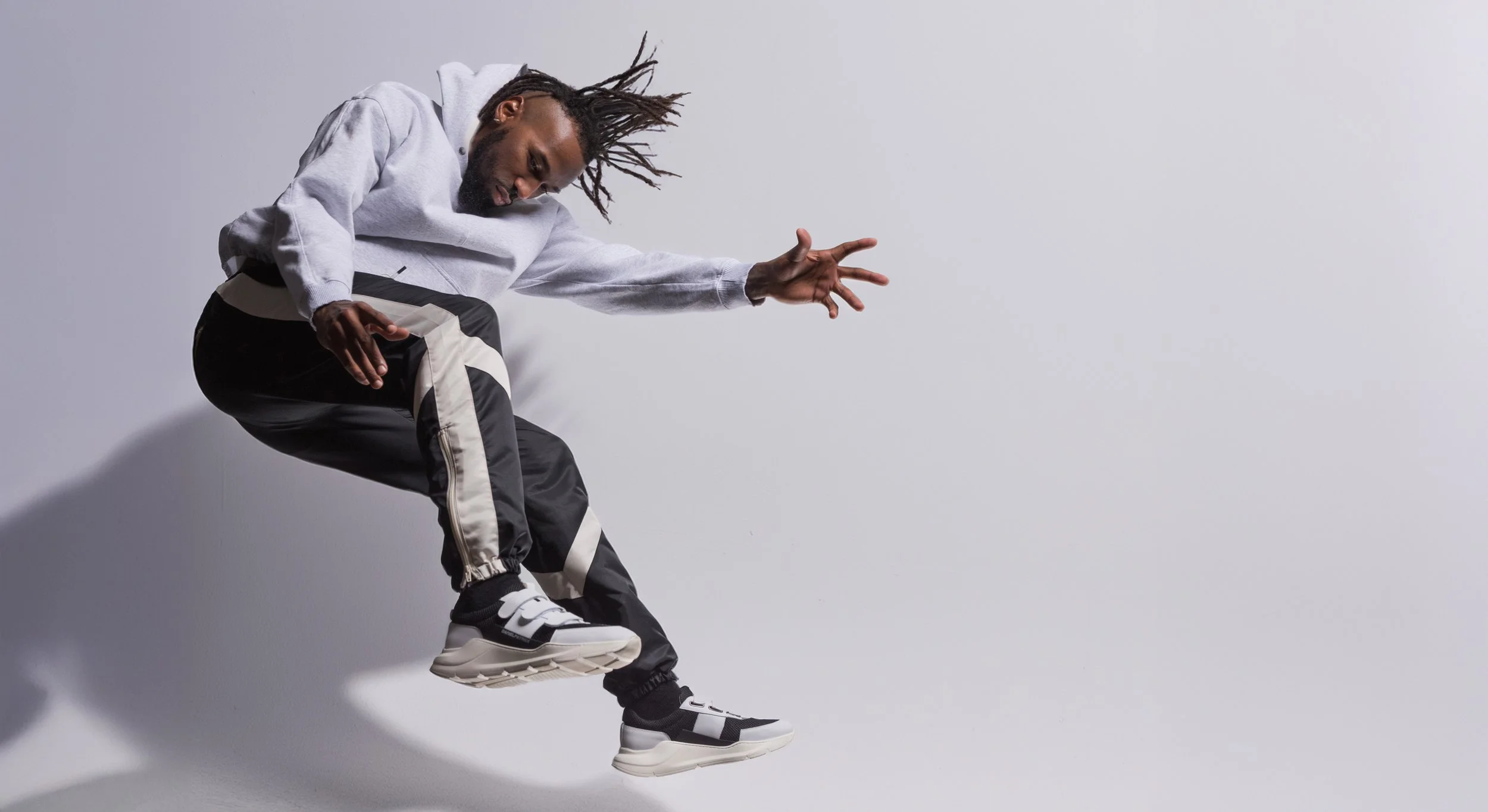 A man wearing a gray hoodie, black and white track pants, and black and white sneakers is mid-air against a gray background, with his head leaning forward and hair flying back.