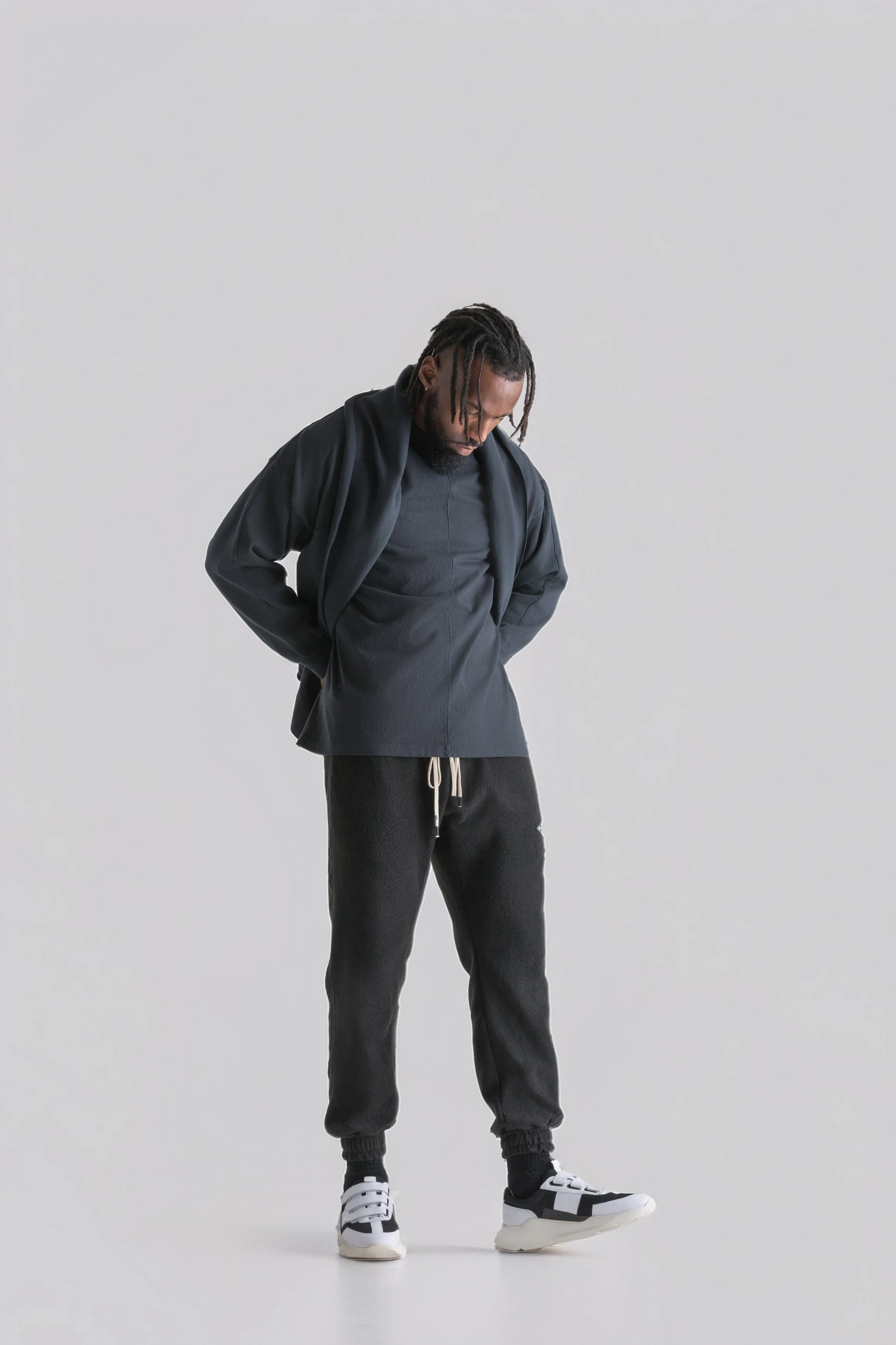 Portrait of a young man dressed in black athletic wear, standing against a plain white background, looking down with hands in pockets.