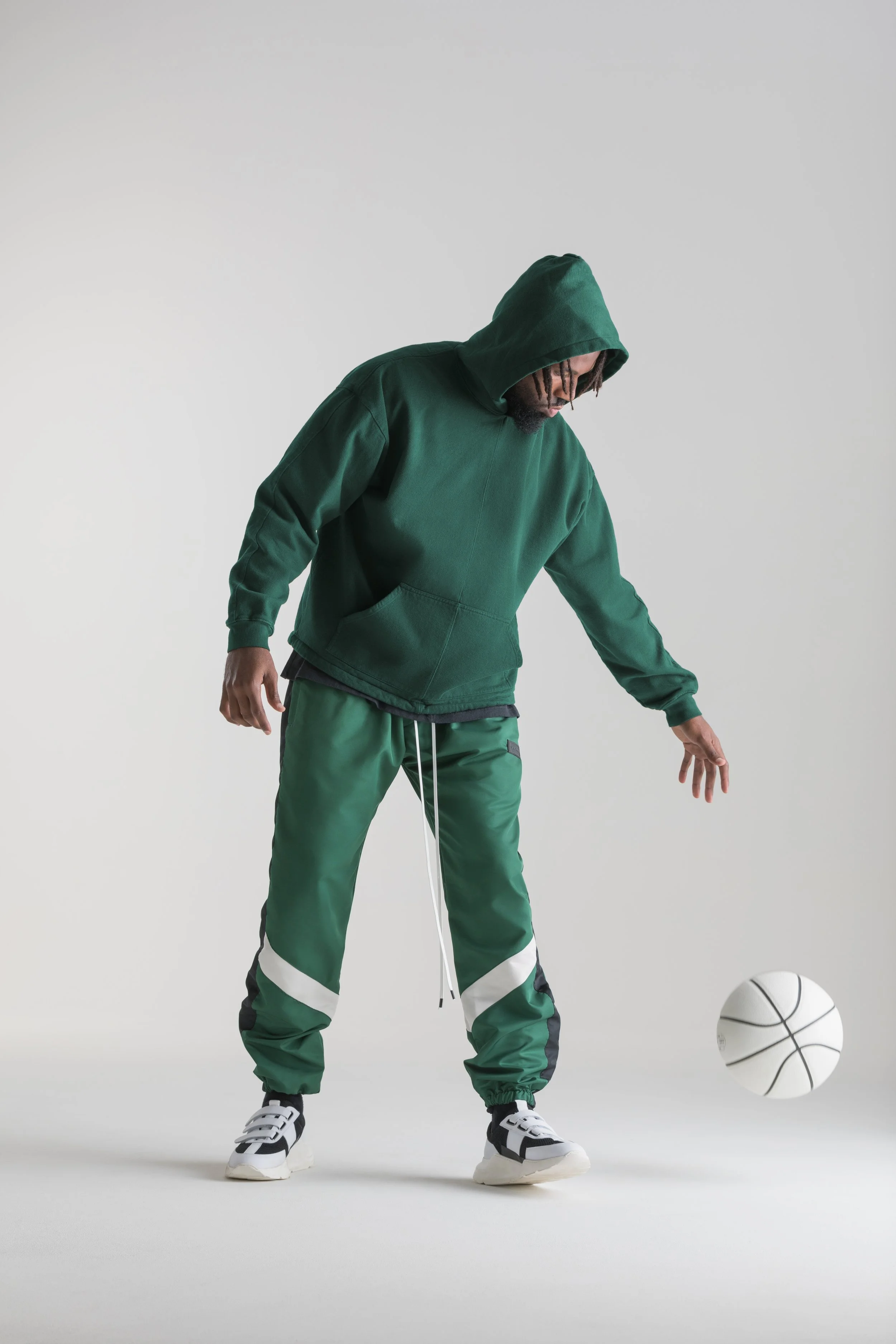 A man dressed in a green hoodie and green joggers is standing on a plain white background, playing with a white basketball that is slightly off the ground.