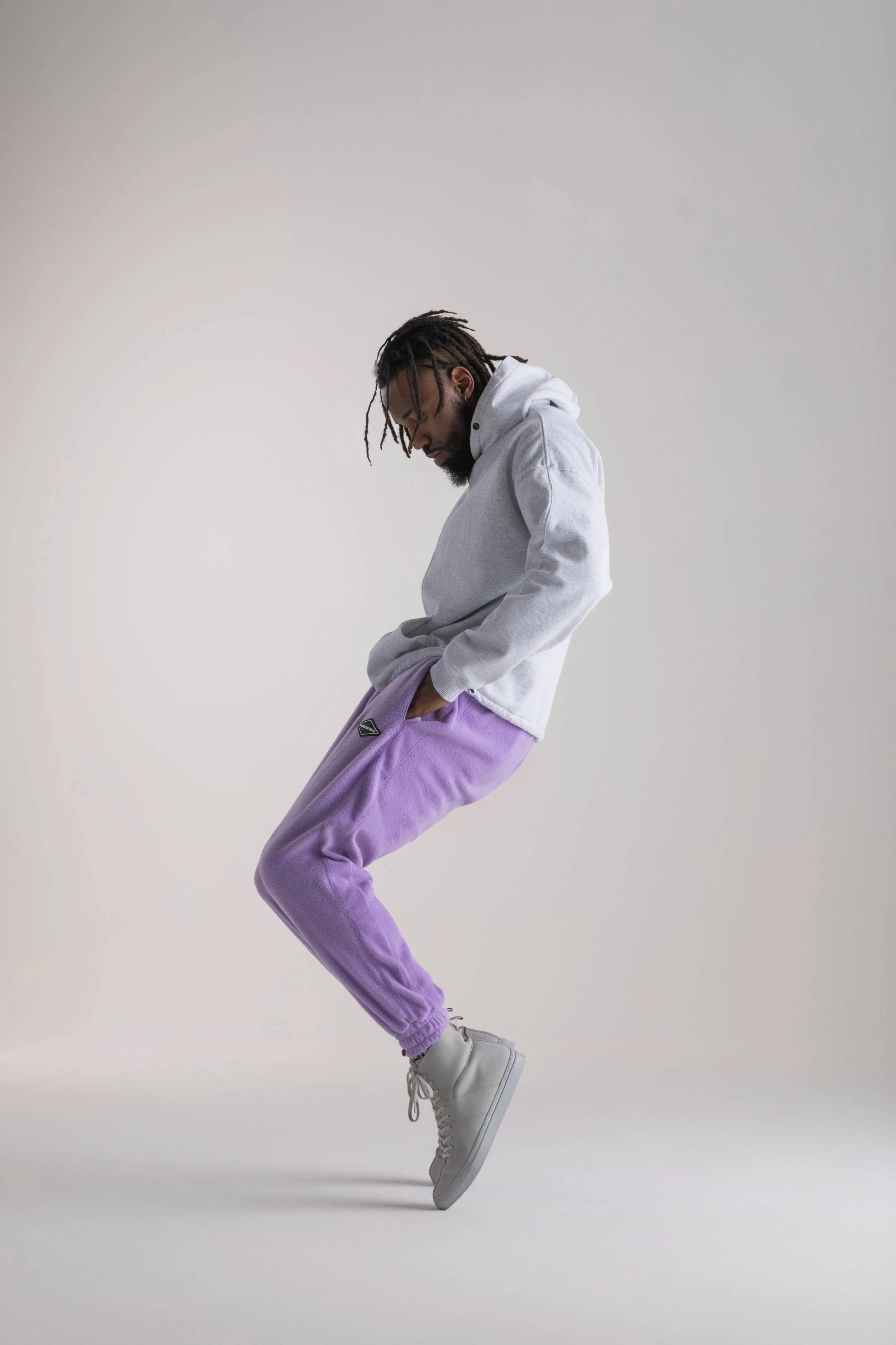 A man jumping in the air against a plain background, wearing a gray hoodie, purple sweatpants, and white high-top sneakers.
