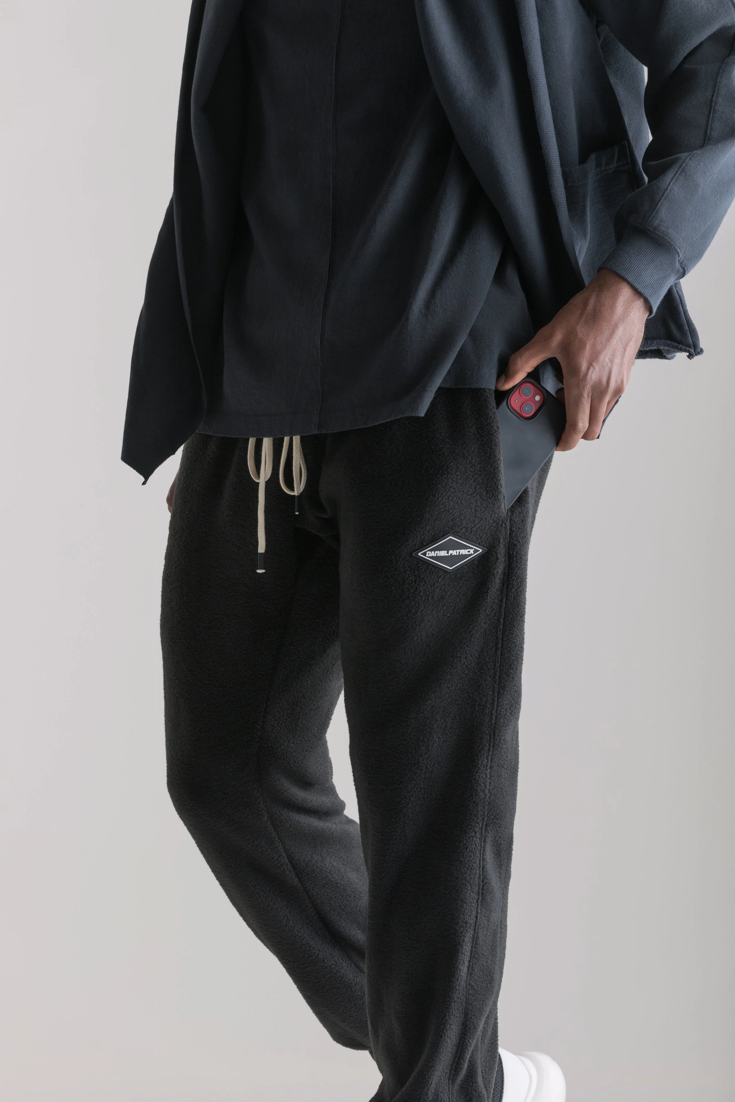 Person wearing black sweatpants with a logo, holding a smartphone in their right hand, with a plain backdrop.