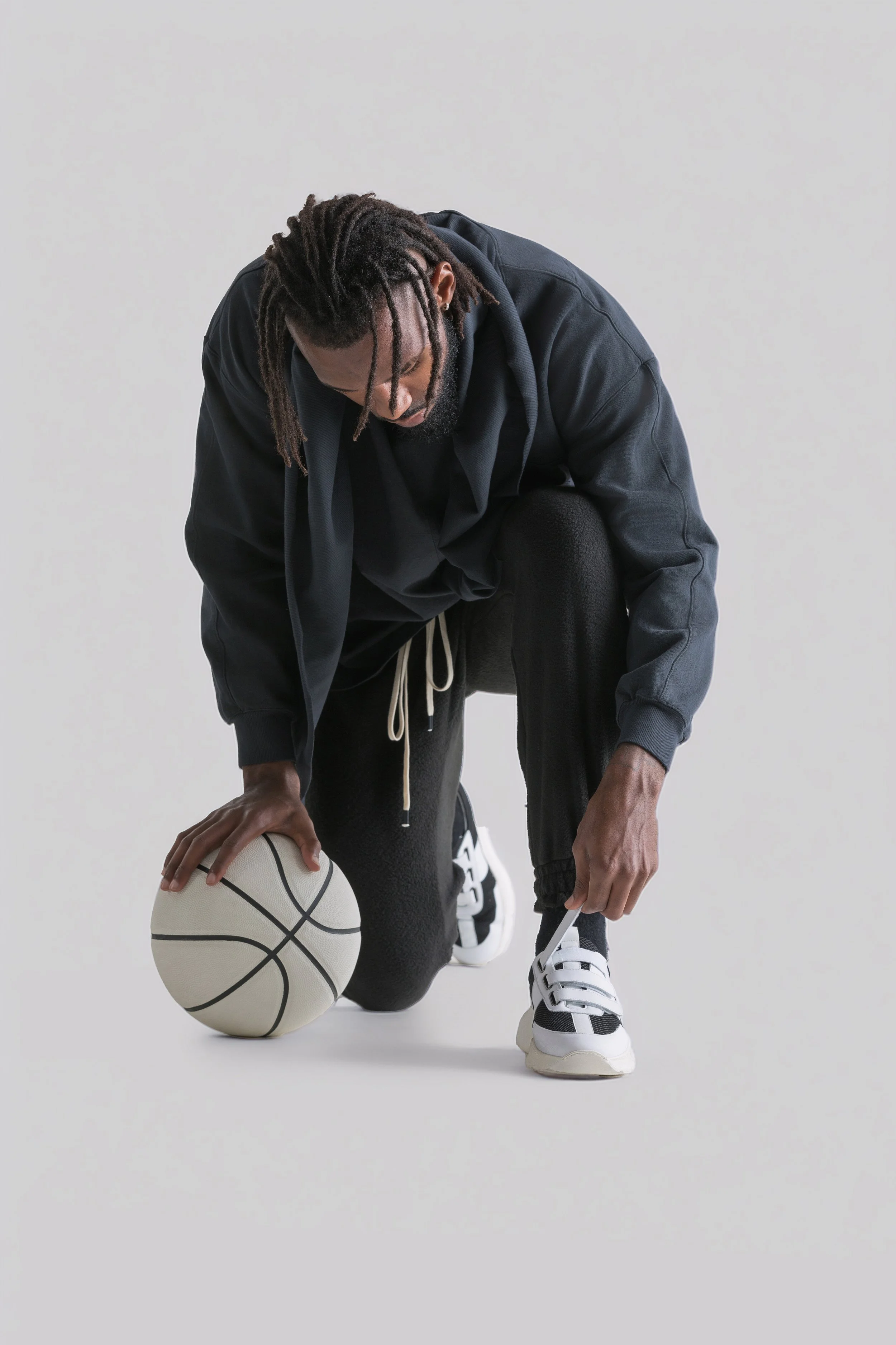 A man wearing black athletic clothing kneels on one knee, holding a stringless basketball with one hand and tying his sneaker with the other, against a plain white background.