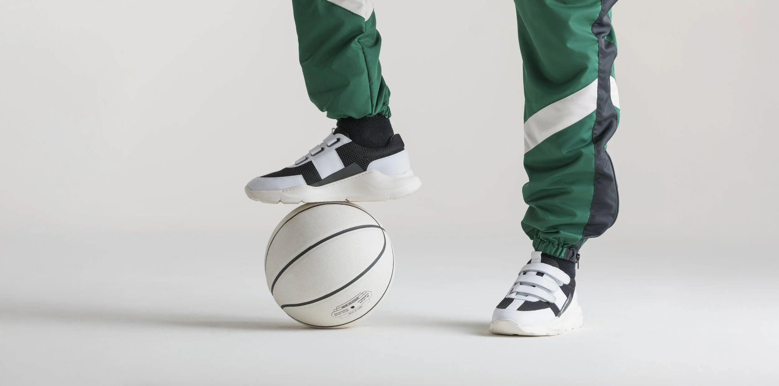 Person wearing black and white sneakers standing on a white basketball, balancing with one foot, against a plain white background.