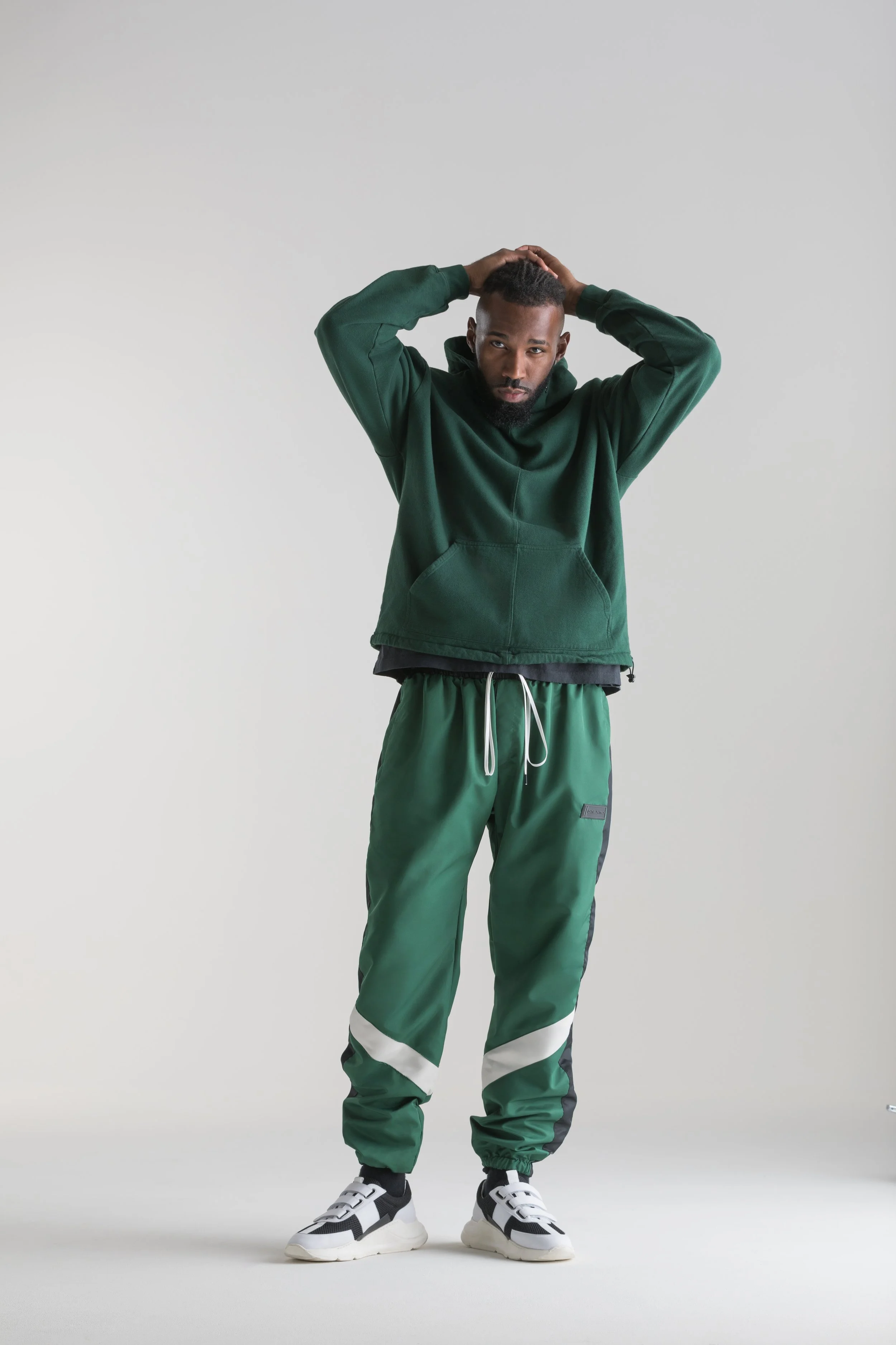 A man standing against a plain background, wearing a green hoodie, green and black jogger pants, and black and white sneakers, with his hands resting on his head.