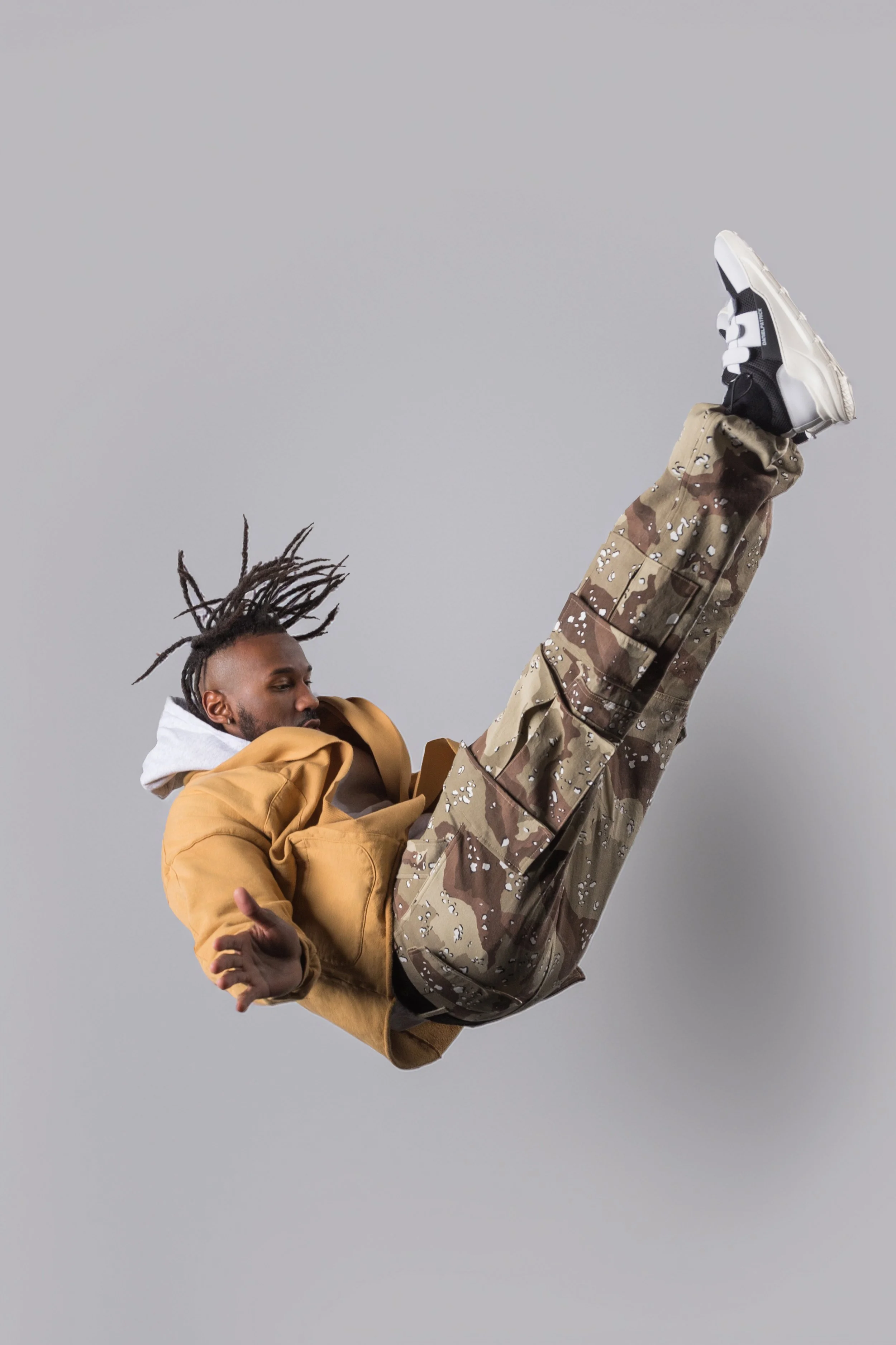 A man with dreadlocks wearing a brown jacket and camouflage pants performs a dance move against a plain gray background.