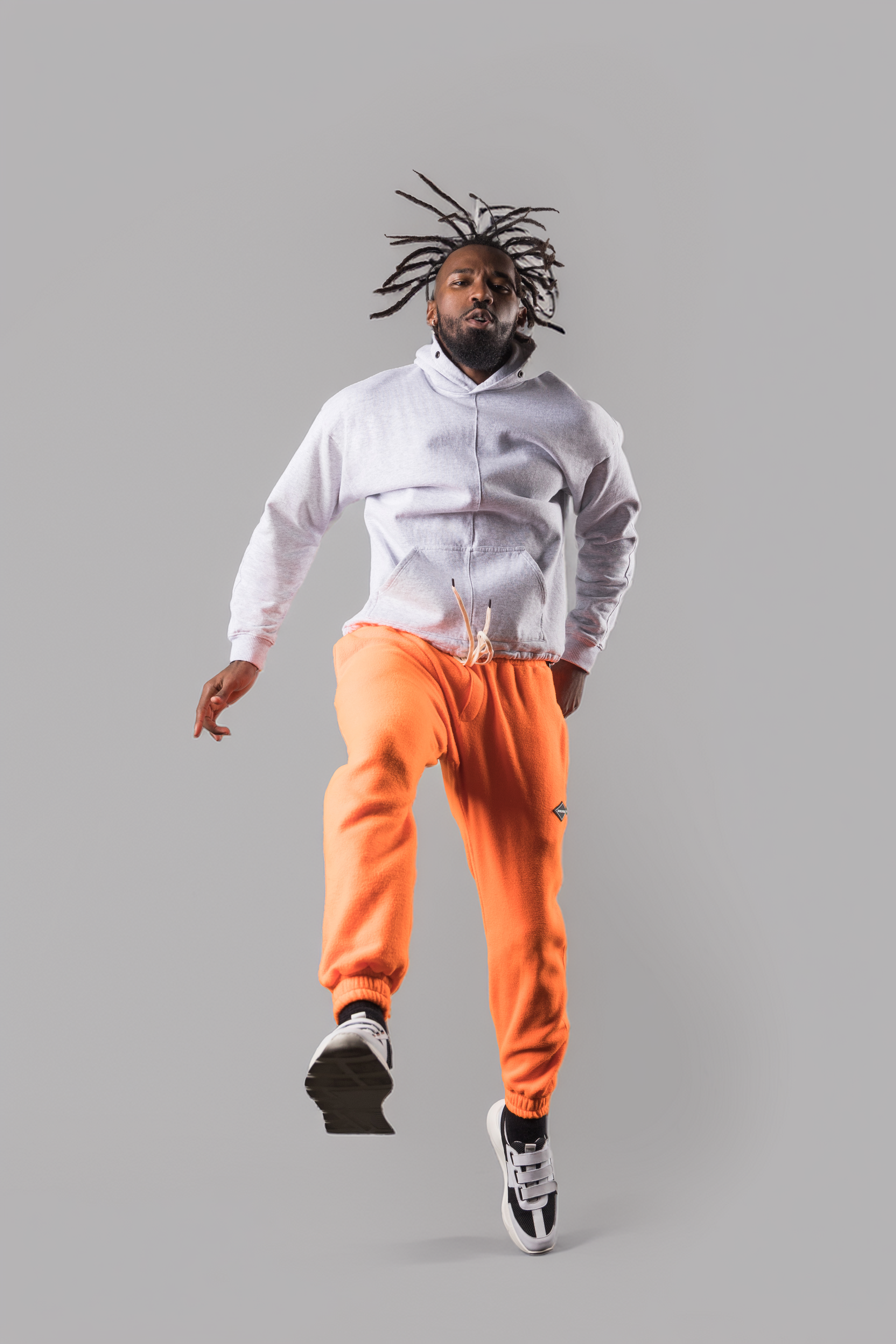 A man with dreadlocks jumping in mid-air against a plain gray background, wearing a gray hoodie, orange sweatpants, and black and white sneakers.