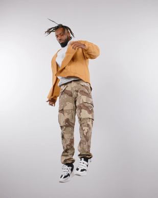 Young man with dreadlocks jumping in a studio with a plain white background, wearing a brown jacket, gray hoodie, camouflage cargo pants, and white sneakers.