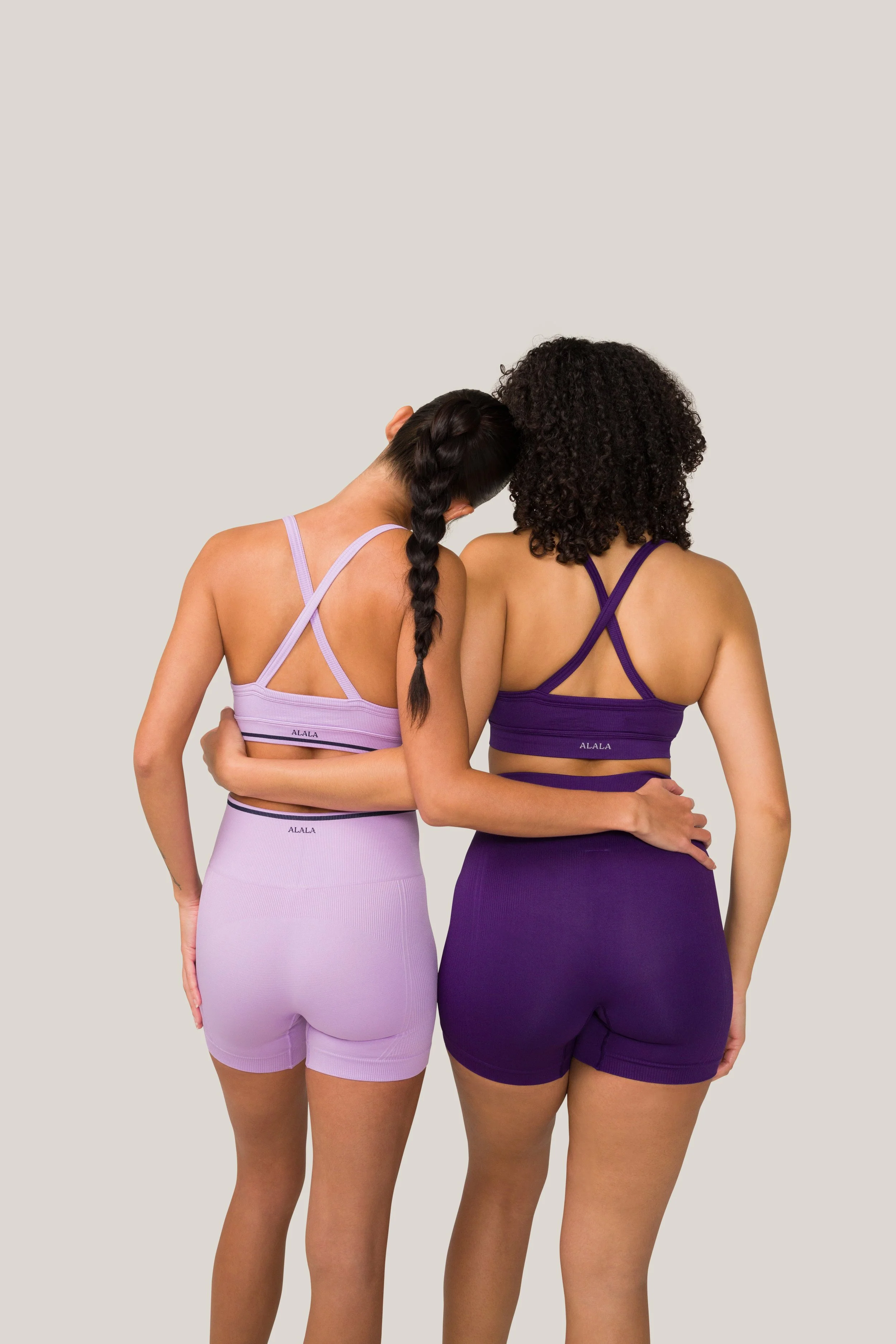 Two women in matching sports bras and shorts, embracing each other against a plain background, one in lavender and the other in dark purple.