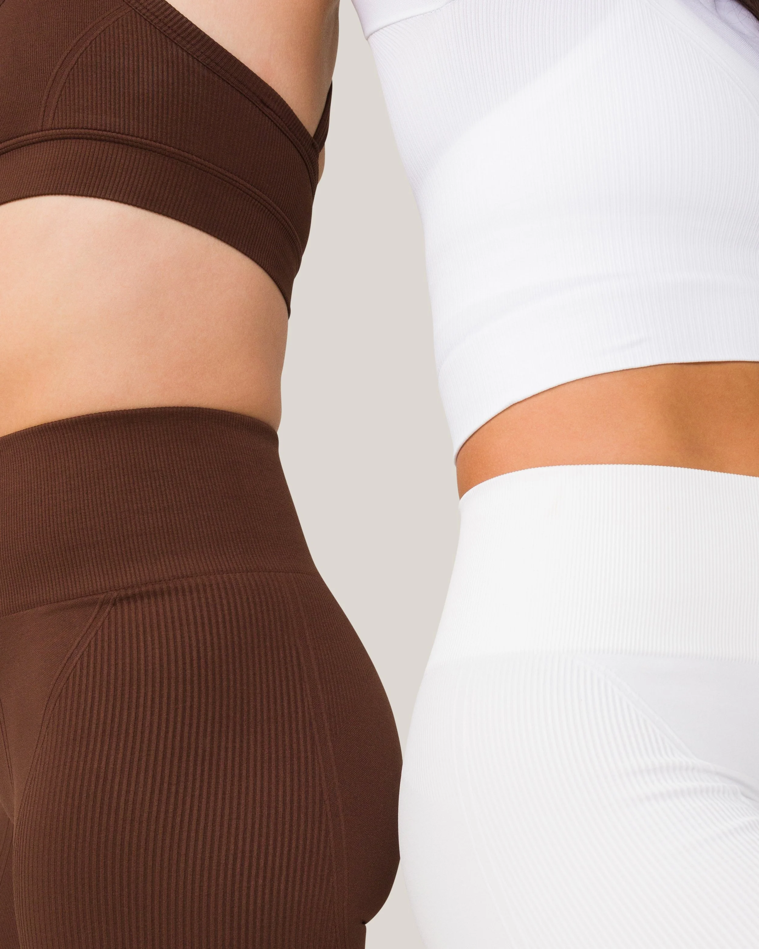 Close-up of two women wearing form-fitting athletic crop tops and high-waisted leggings, one in brown and the other in white, standing side by side.
