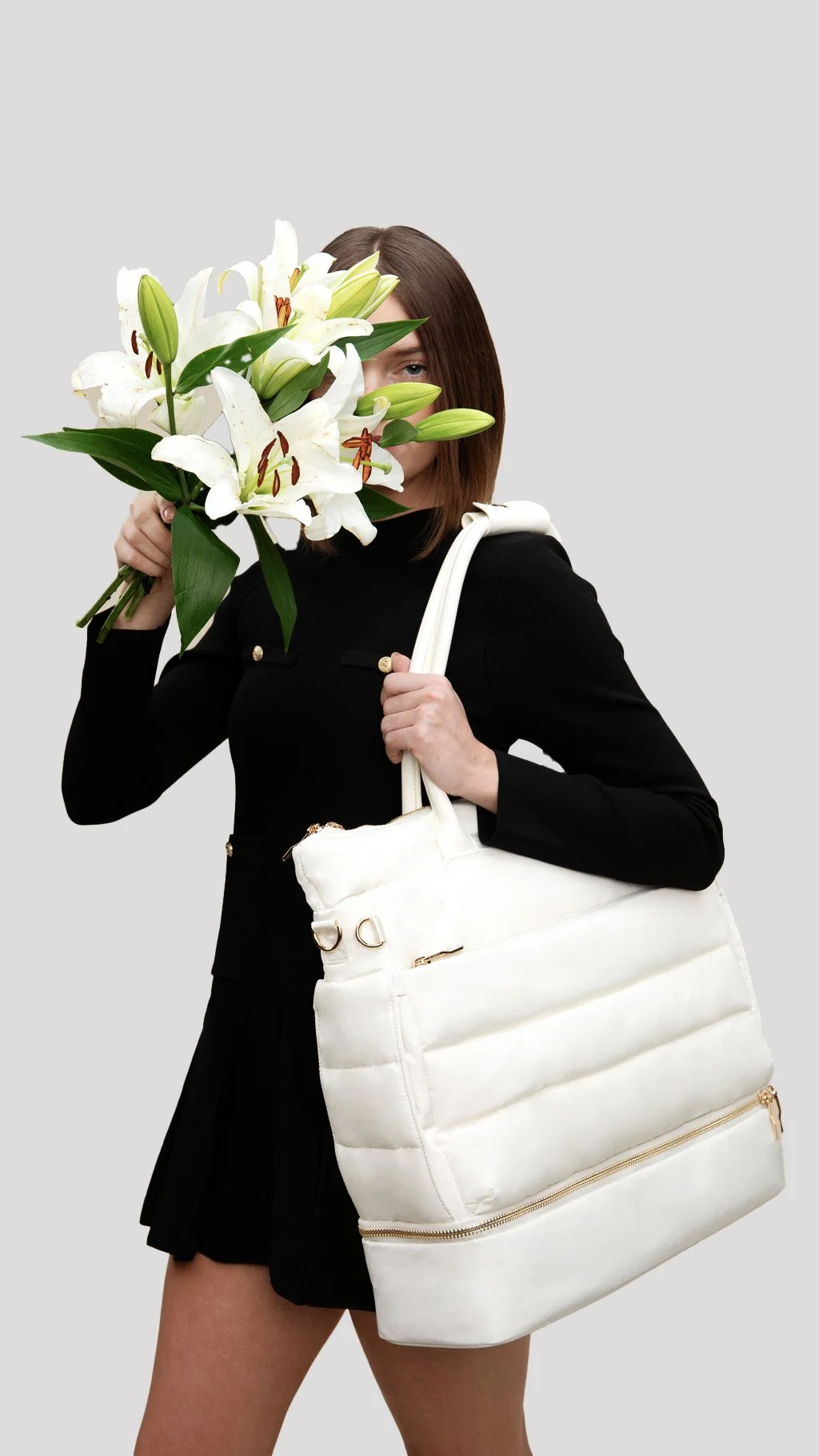 A woman with shoulder-length brown hair holds a bouquet of white lilies in front of her face, partially covering her features. She is dressed in a black blazer with gold buttons and carries a large white duffle bag over her shoulder. The background is plain and light grey.