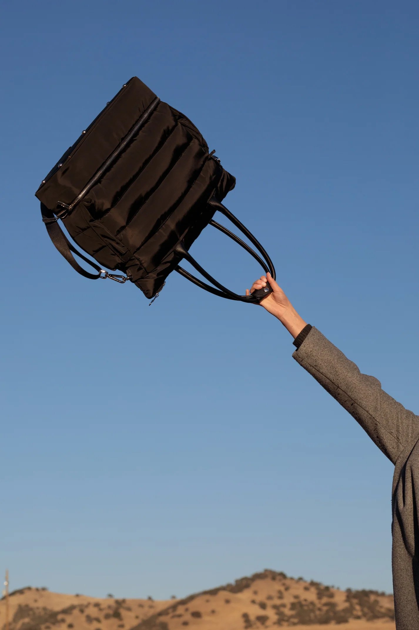 A person holding a black backpack in the air outdoors against a blue sky with hills in the background.