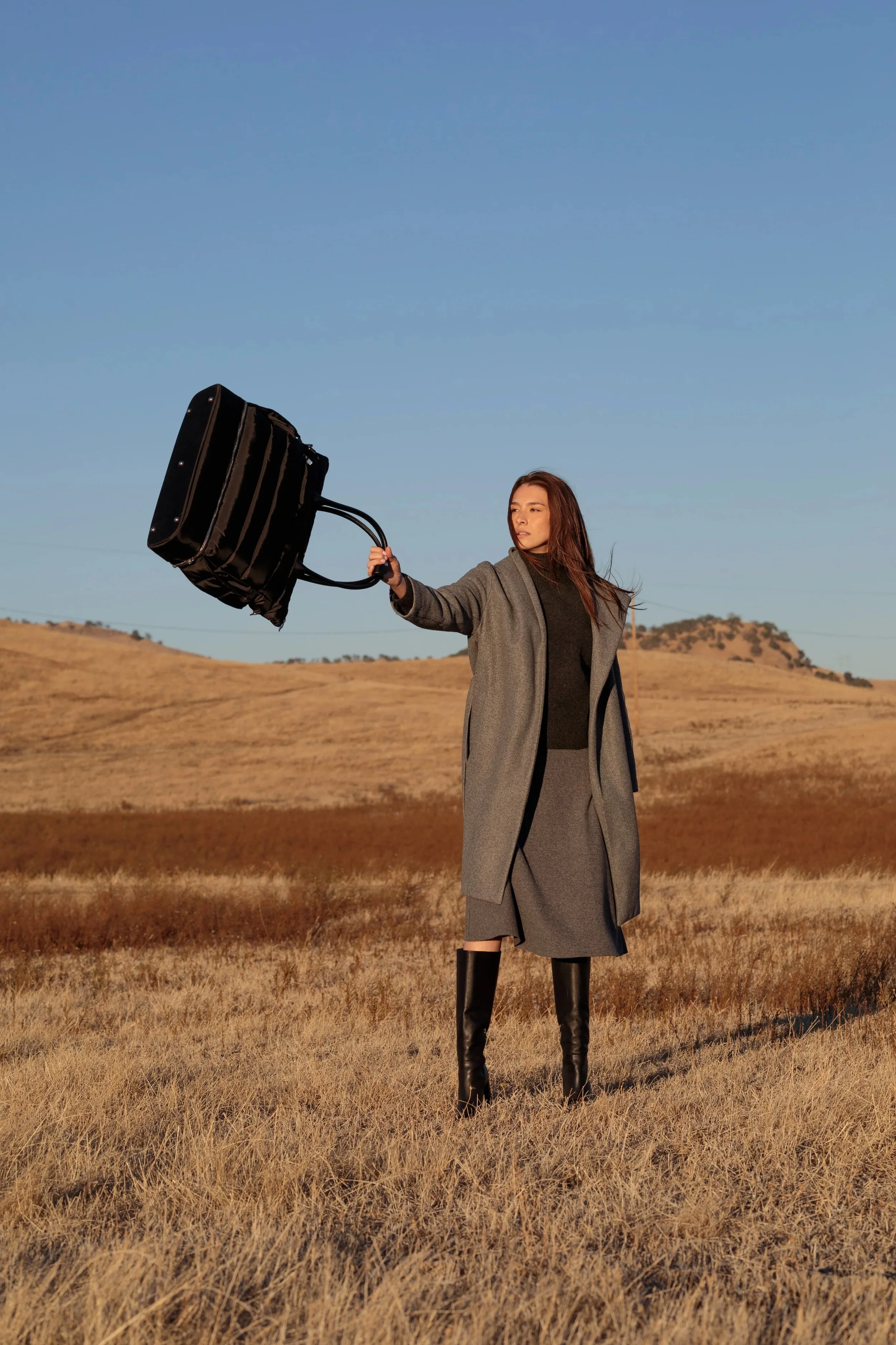 A woman in a gray coat and knee-high black boots stands in a field, holding a rolling suitcase in her right hand, against a clear blue sky and rolling hills.