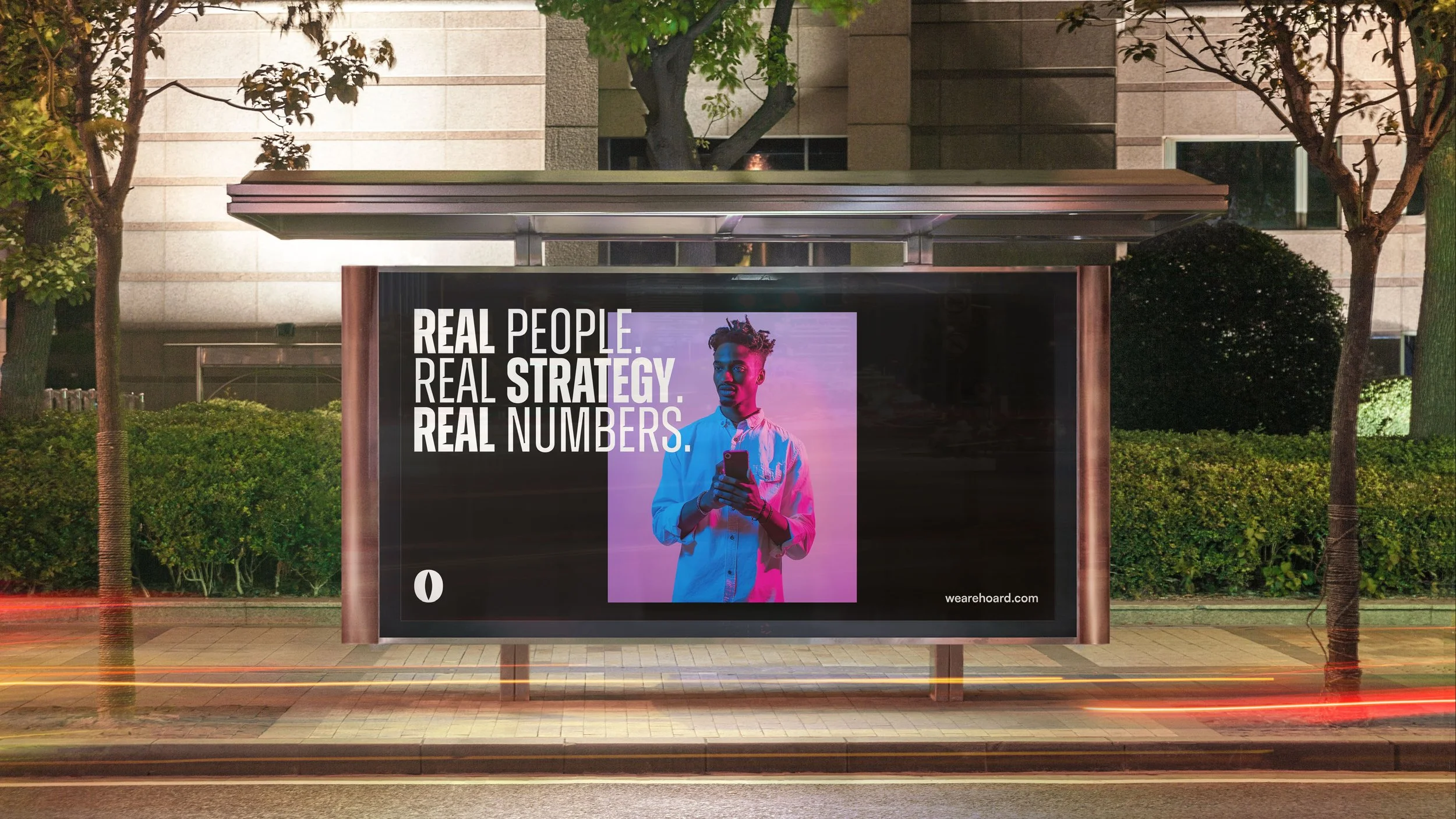 An outdoor bus stop with a digital advertisement showing a young man holding a phone, with the text "Real People. Real Strategy. Real Numbers." in bold white and purple letters, against a pink and purple background with trees and a building in the background.
