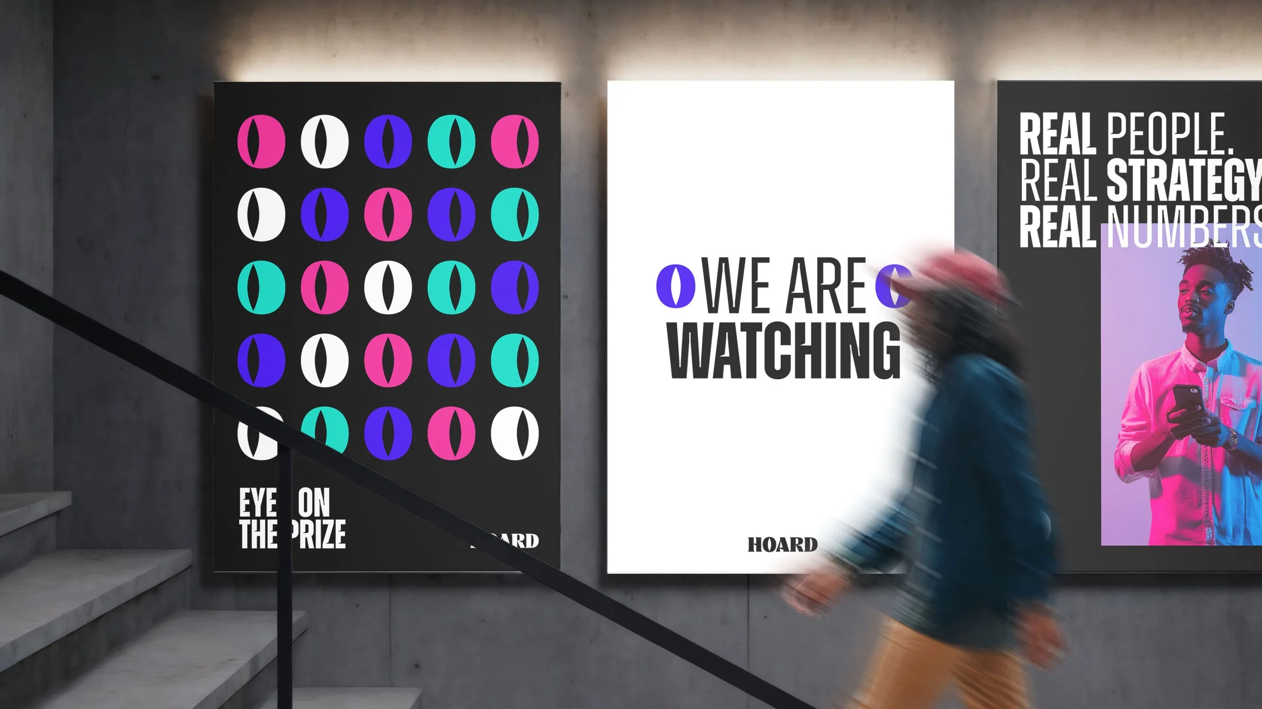 Three posters on a gray concrete wall in an art gallery or museum, with a person walking past them. The left poster features colorful eye-like shapes on a black background with the text 'EYE ON THE PRIZE.' The middle white poster displays the text 'WE ARE WATCHING' with purple eye symbols. The right poster has a black background with white text reading 'REAL PEOPLE. REAL STRATEGY. REAL NUMBERS.'
