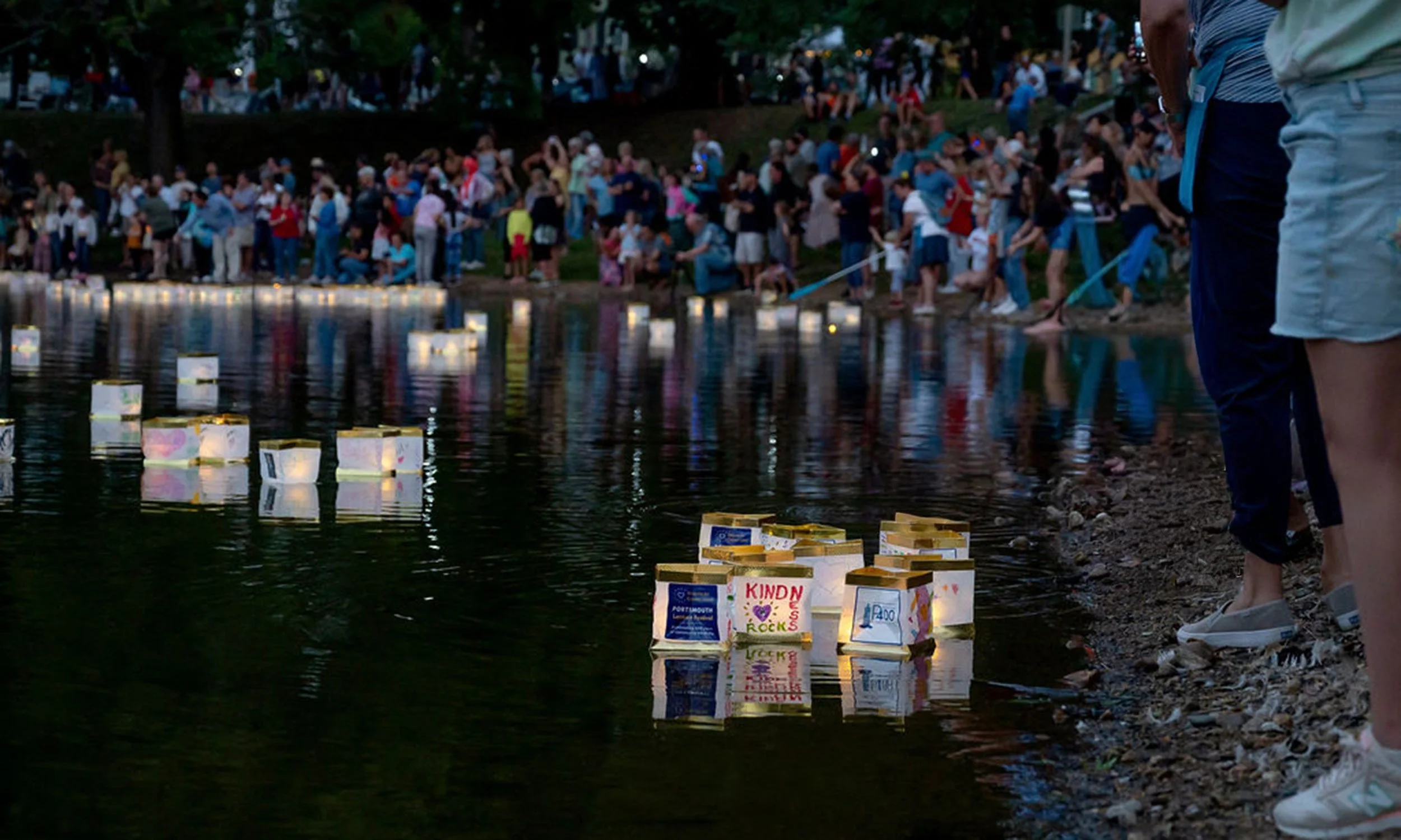 People gathered along a riverbank during dusk, releasing floating lanterns with colorful designs onto the water.