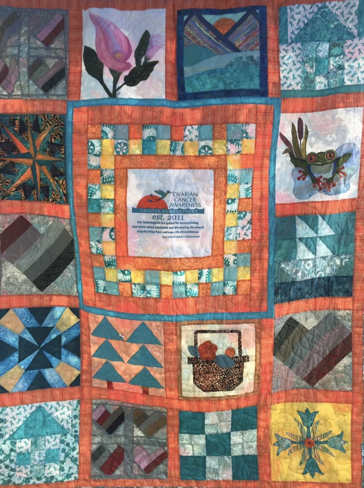 A quilt with multiple square patches, including a central patch with the Ovarian Cancer Awareness logo and text, surrounded by various colorful patterned fabric squares, including flowers, animals, and geometric designs.