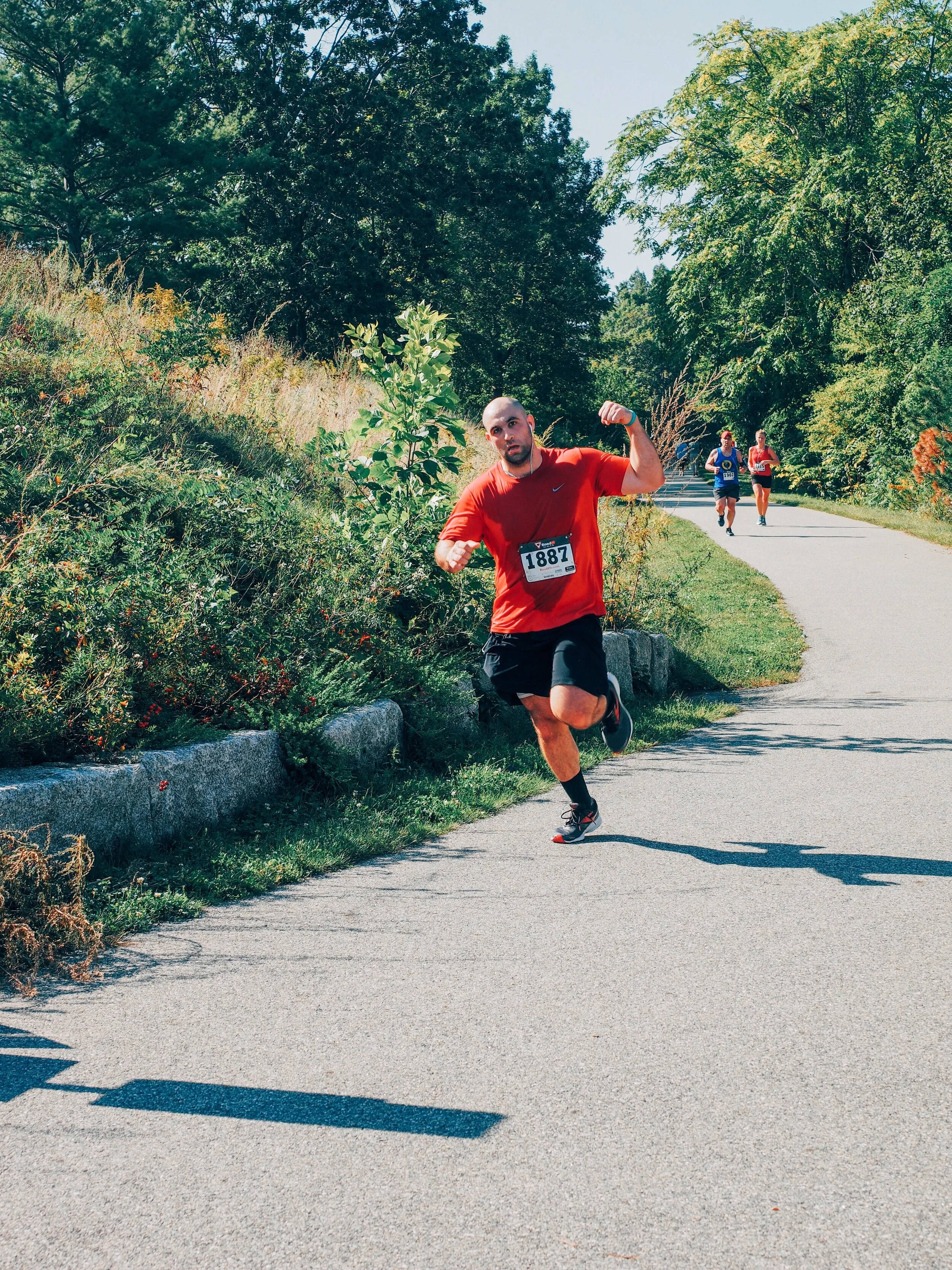 A man in a red shirt and black shorts running on a paved trail during a race, with a race number 1887, surrounded by green trees and bushes, with two other runners in the background.