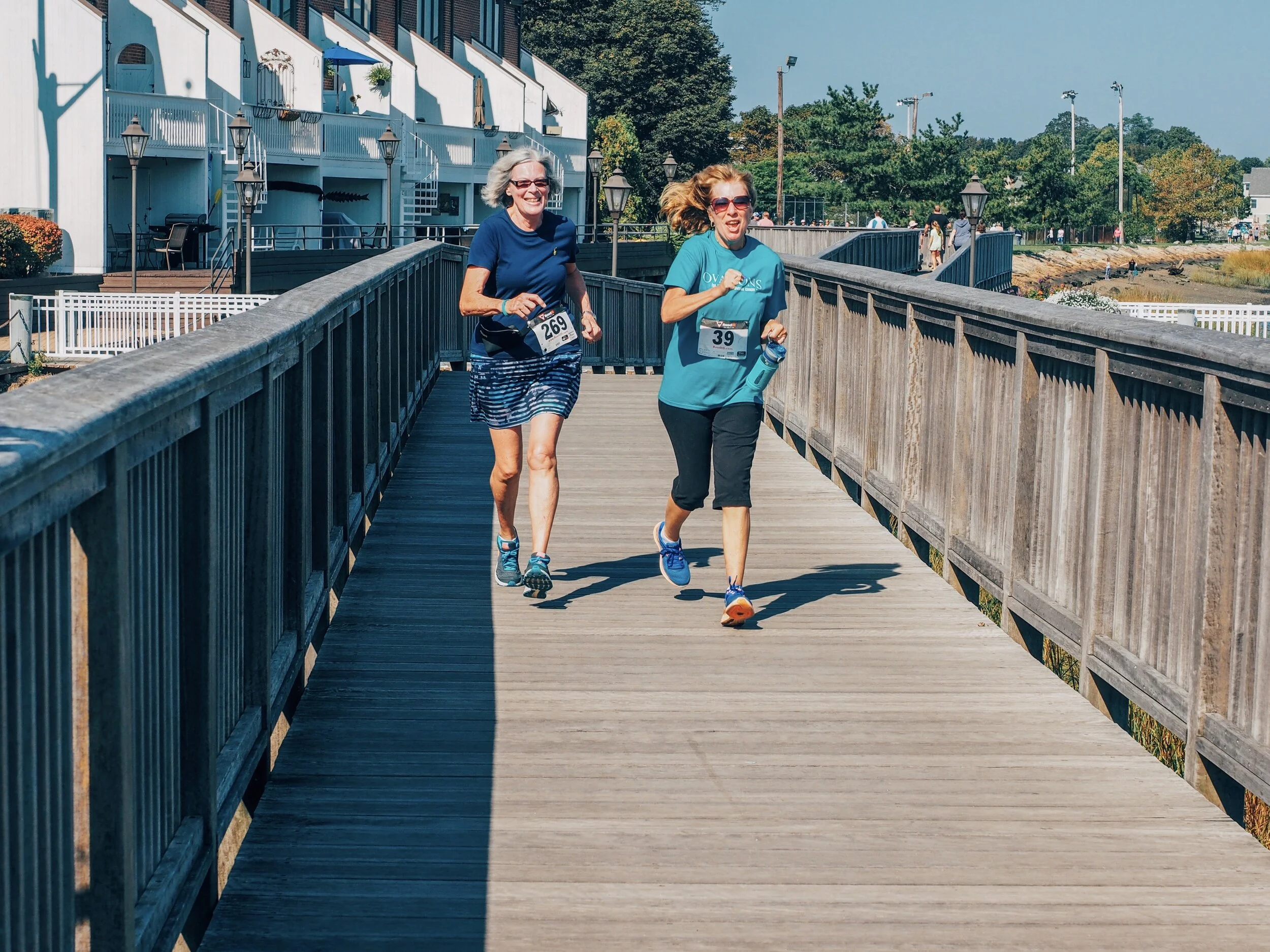 Two women running on a wooden bridge during a race, wearing numbered bibs, with trees and buildings in the background on a sunny day.
