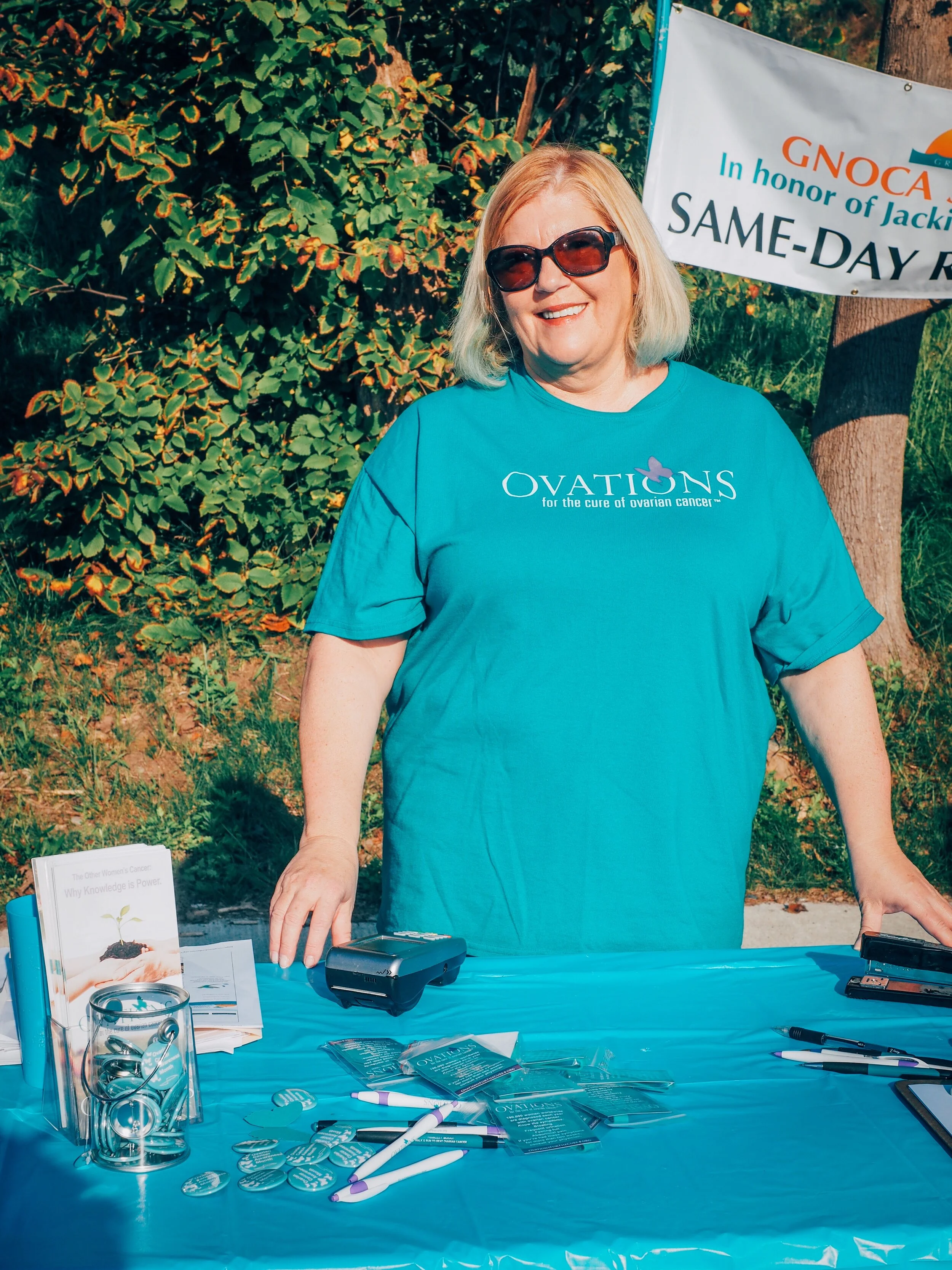 Smiling woman with blonde hair and sunglasses standing behind an information table at an outdoor ovarian cancer awareness event, wearing a blue OVATIONS t-shirt with ovarian cancer awareness ribbon. The table has brochures, pens, buttons, and a stamp