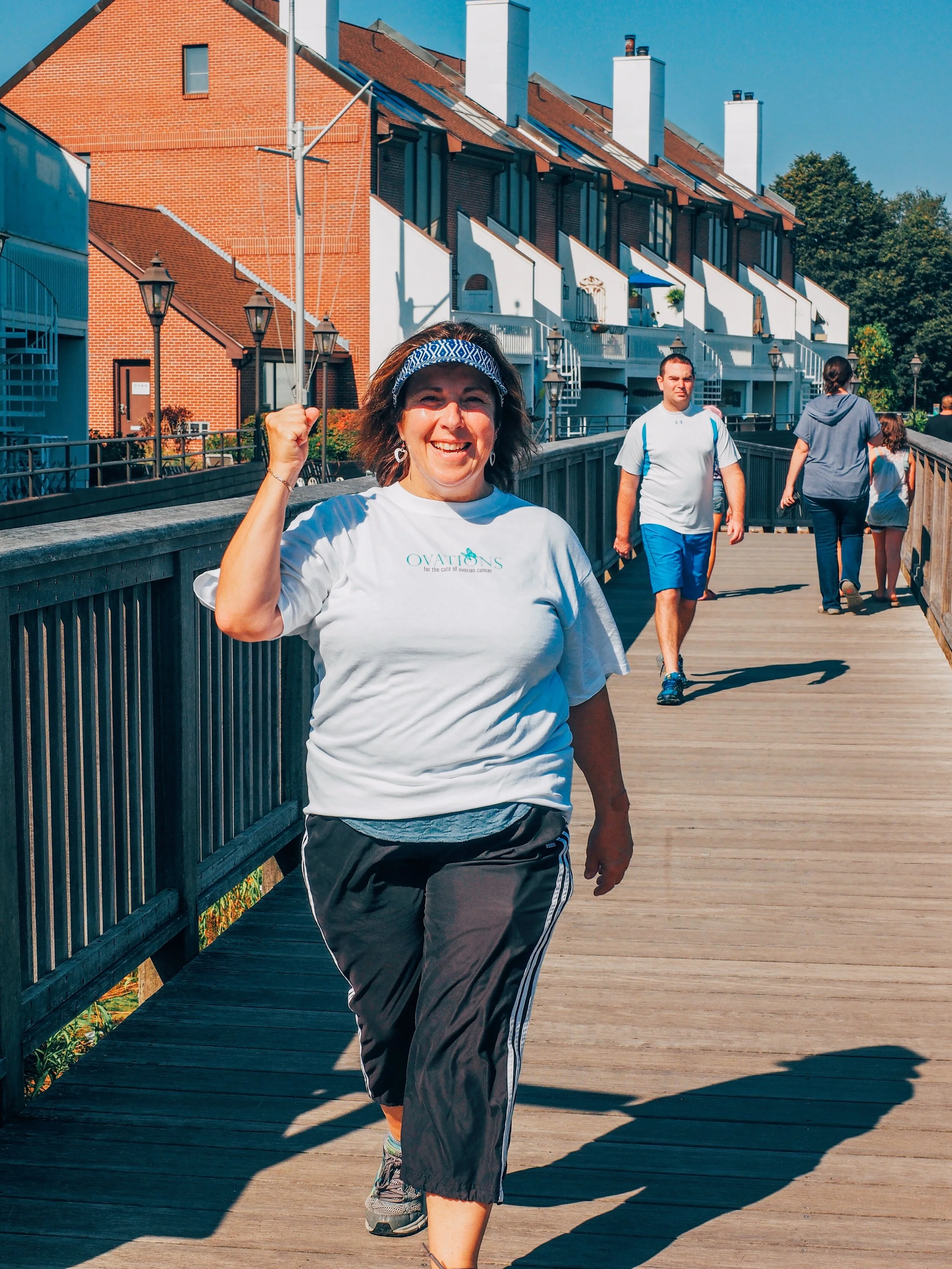 Smiling woman jogging on a wooden bridge with other people walking behind her, in a neighborhood with brick and modern houses under a blue sky.