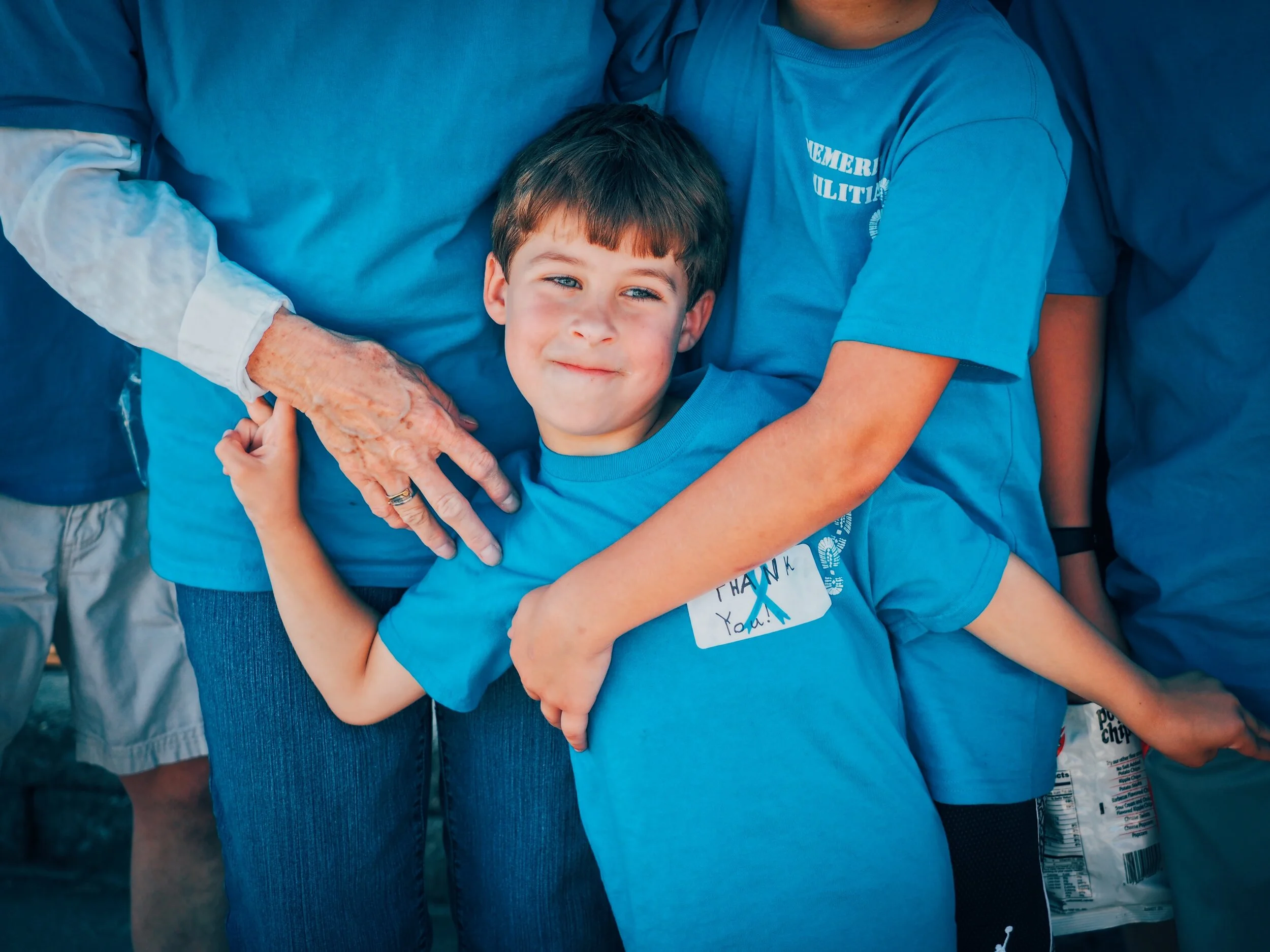 A young boy wearing a blue t-shirt with a thank you note and ribbon, being embraced by two adults in matching blue shirts, one with an outstretched hand on his arm, at an outdoor event.