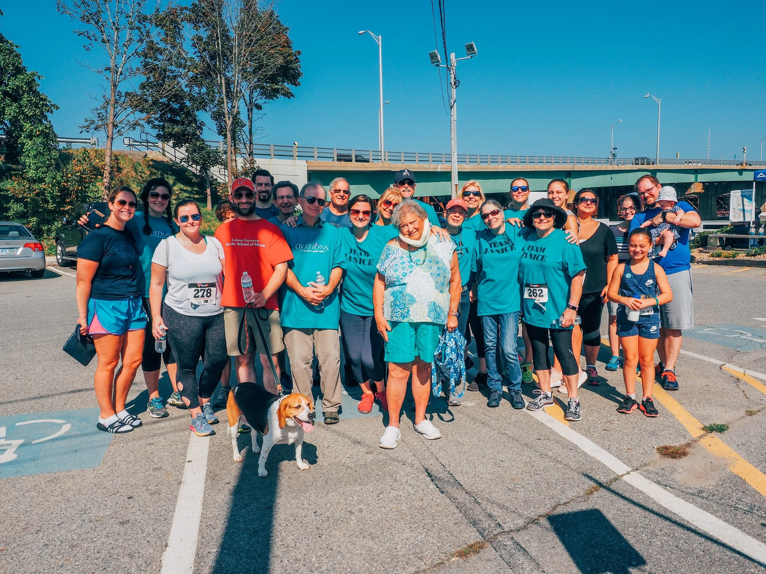 Group of people gathered outdoors in a parking lot on a sunny day, some wearing matching blue shirts, with a woman elderly woman and a beagle dog in the front.