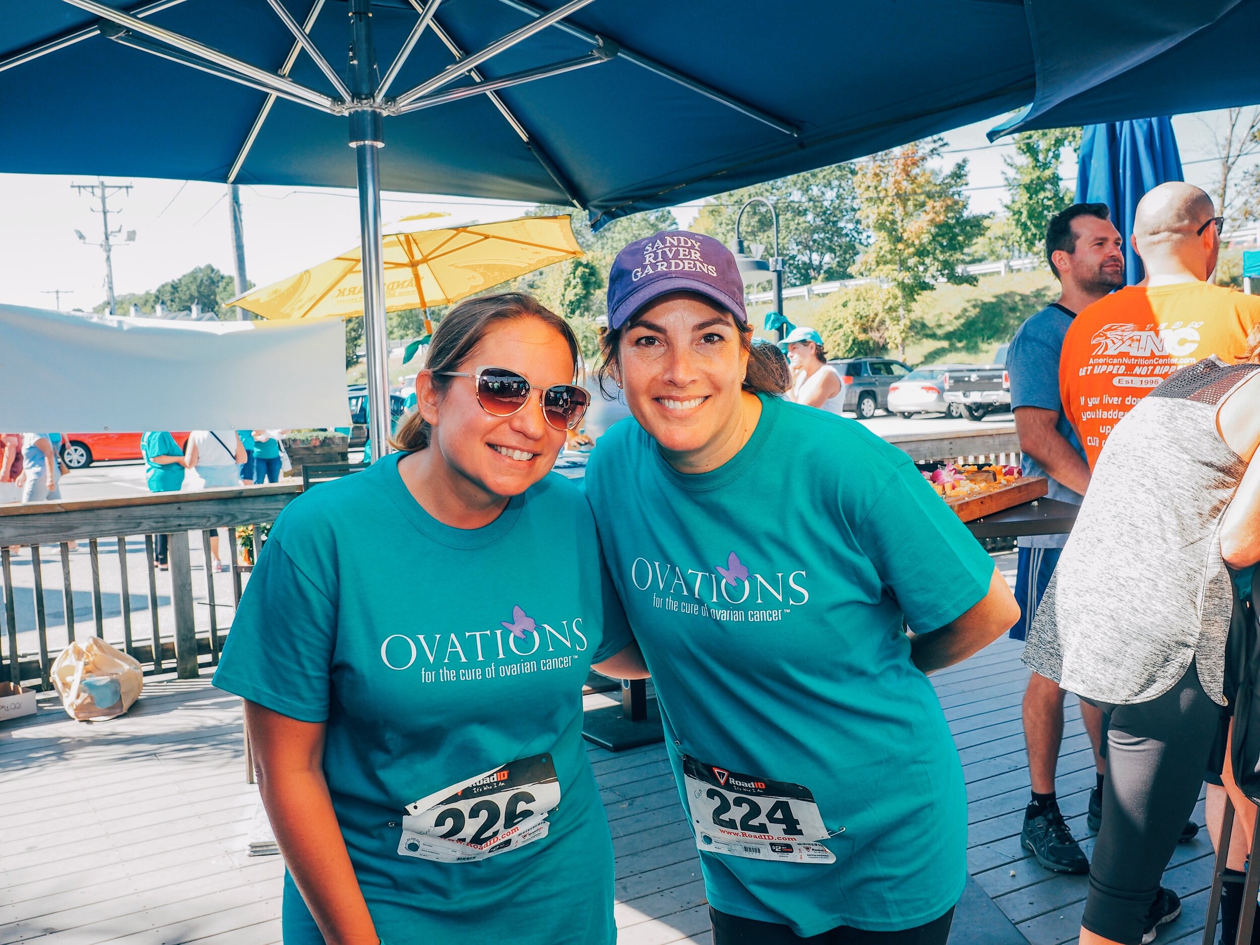 Two women wearing teal shirts with a purple ribbon logo and the text "Ovation for the cure of ovarian cancer" stand together at a daytime outdoor event. One woman wears sunglasses and a race bib numbered 226, and the other woman wears a purple cap an