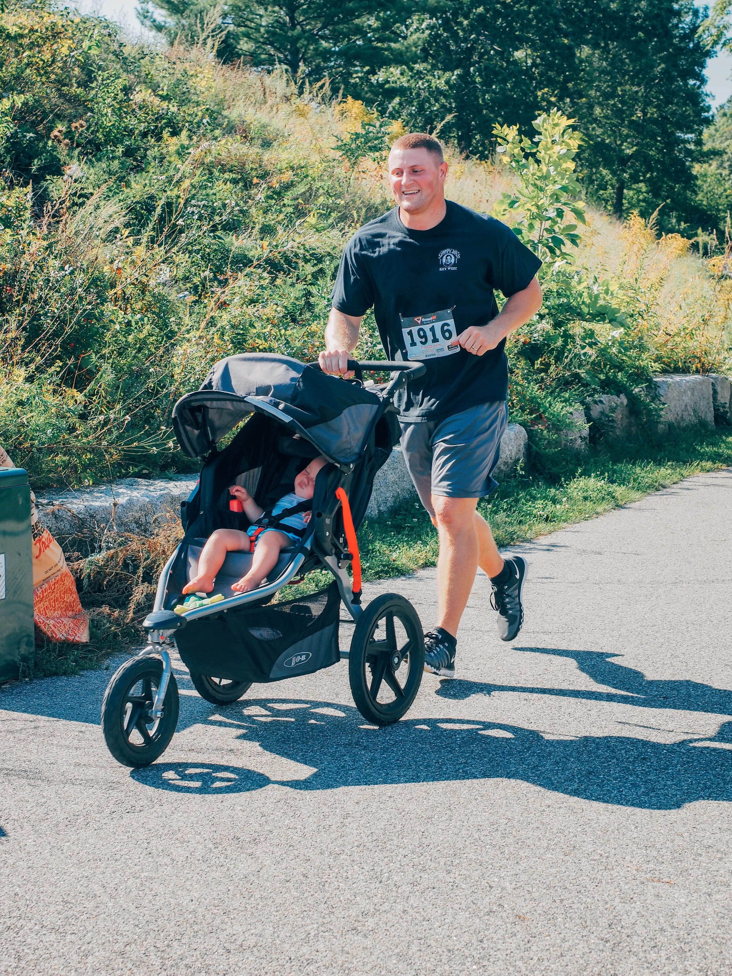 A man jogging with a young child in a stroller along a paved path surrounded by greenery on a sunny day.