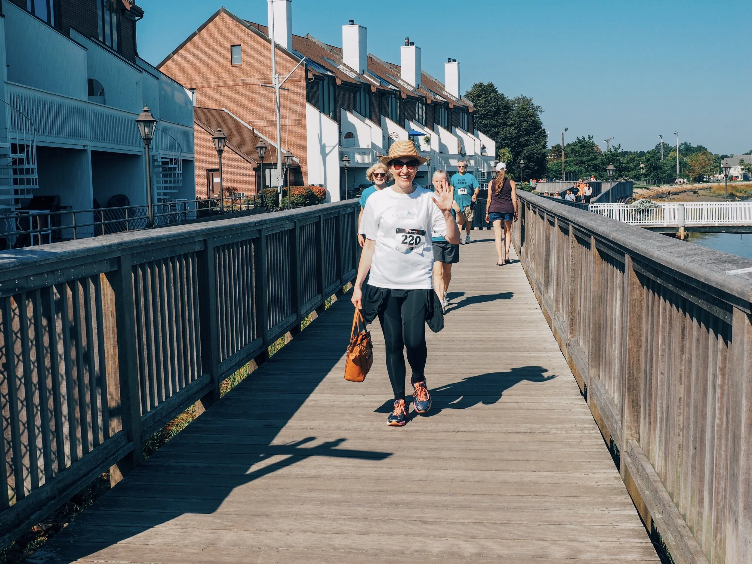 A woman in a white shirt with a race bib number 220, wearing a sunhat, sunglasses, black leggings, and running shoes, is waving and smiling while walking on a wooden pier with other people participating in a running event on a sunny day.