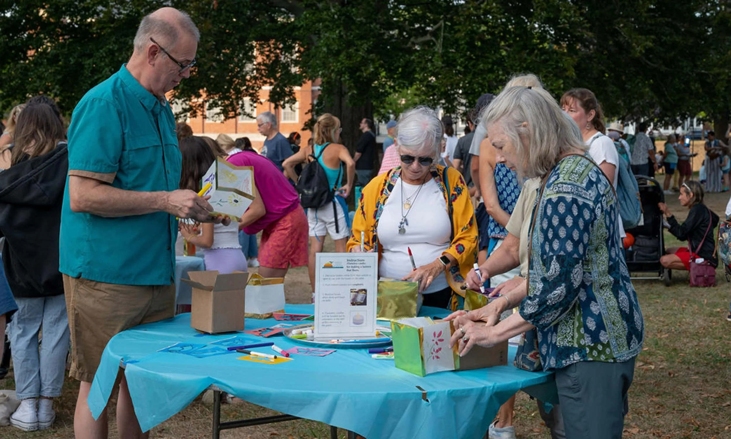 People gather around a table at an outdoor event or festival, engaging in activities like signing papers or filling out forms. The scene is set in a park with large trees and many attendees in casual summer clothing.