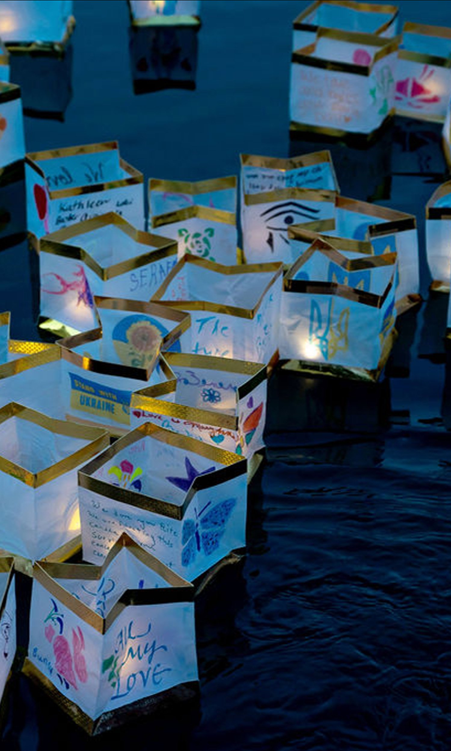 Floating paper lanterns adorned with colorful drawings and handwritten messages are on a body of water, illuminated from within.