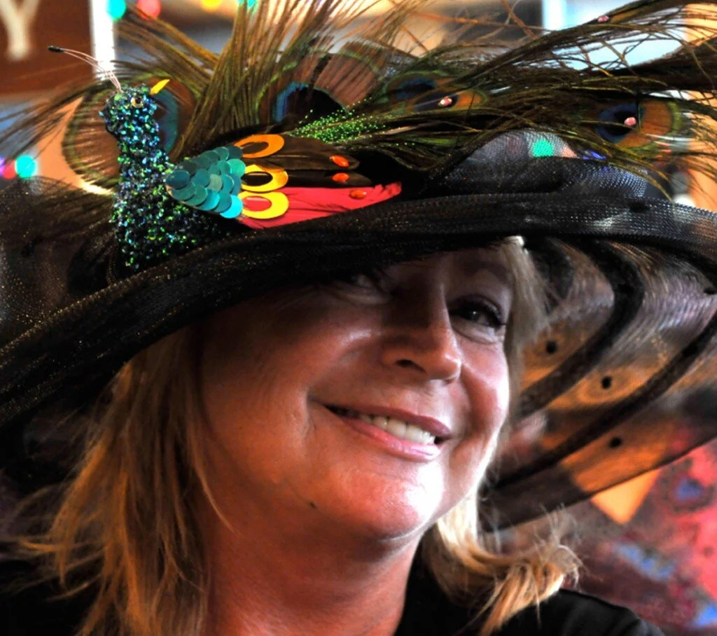 A woman smiling, wearing an elaborate black hat decorated with peacock feathers, sequins, and colorful accents.