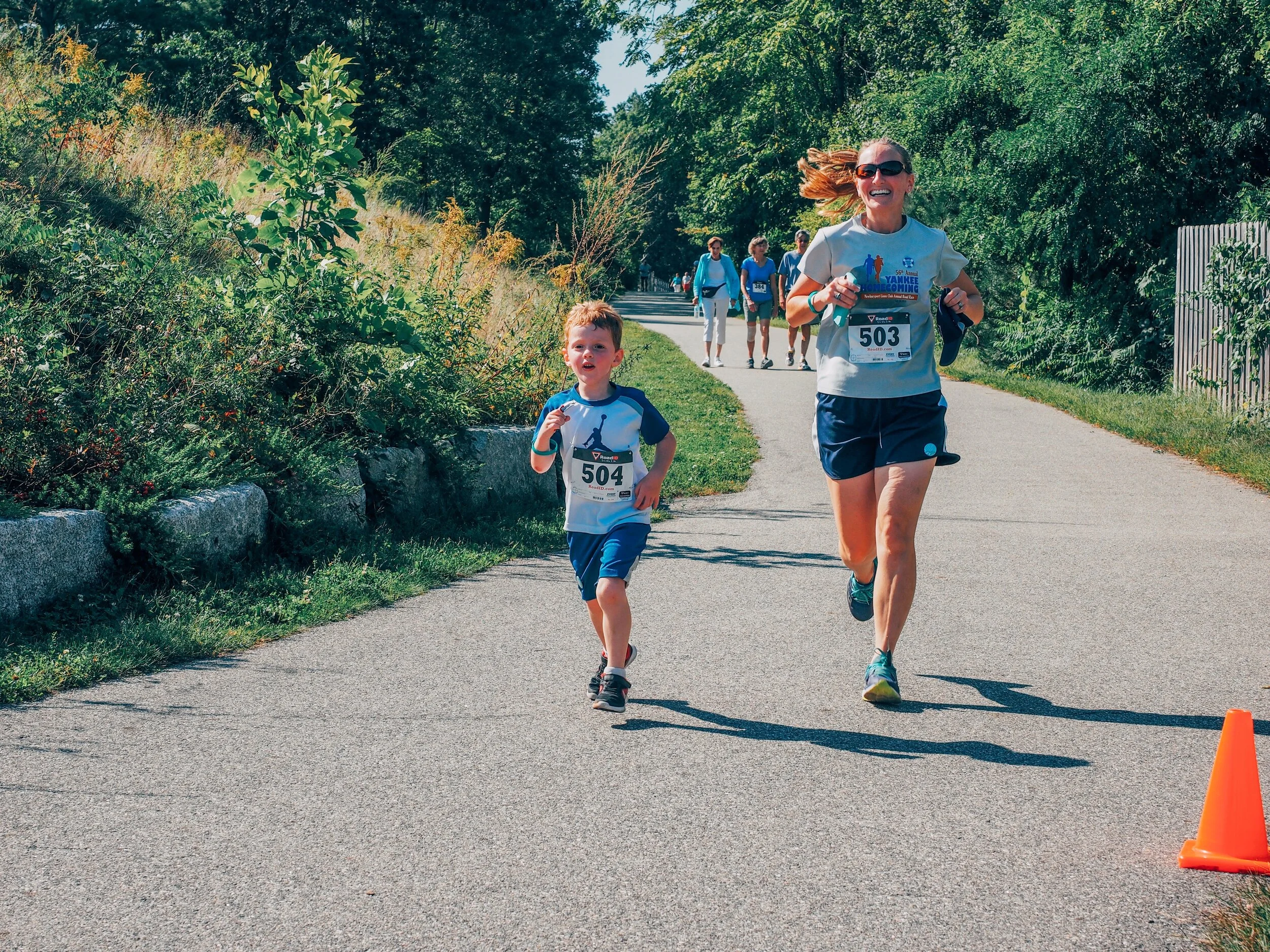 Woman and young boy running outdoors during a race with other runners in the background, on a paved pathway surrounded by greenery and orange traffic cone.