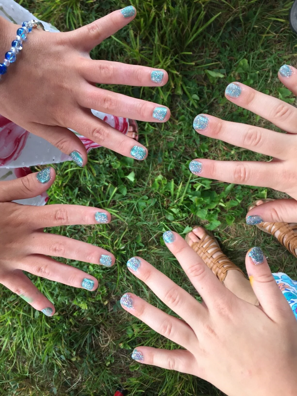 Multiple hands with glittery light blue nail polish, some wearing jewelry, on grass.