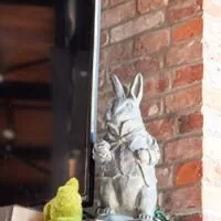 Small gray bunny in a glass jar next to a yellow object, in front of a brick wall.