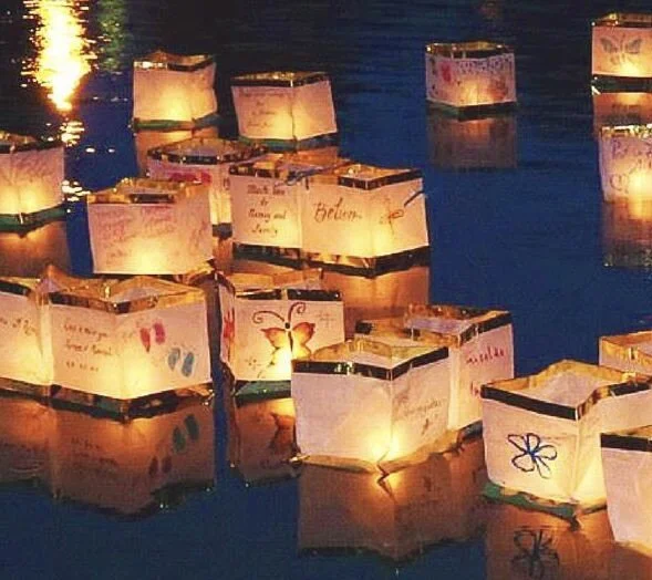 Paper lanterns floating on water at night, some with colorful drawings and writings.