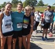 Three young women in athletic clothing standing outdoors at a running event, with other participants in the background.